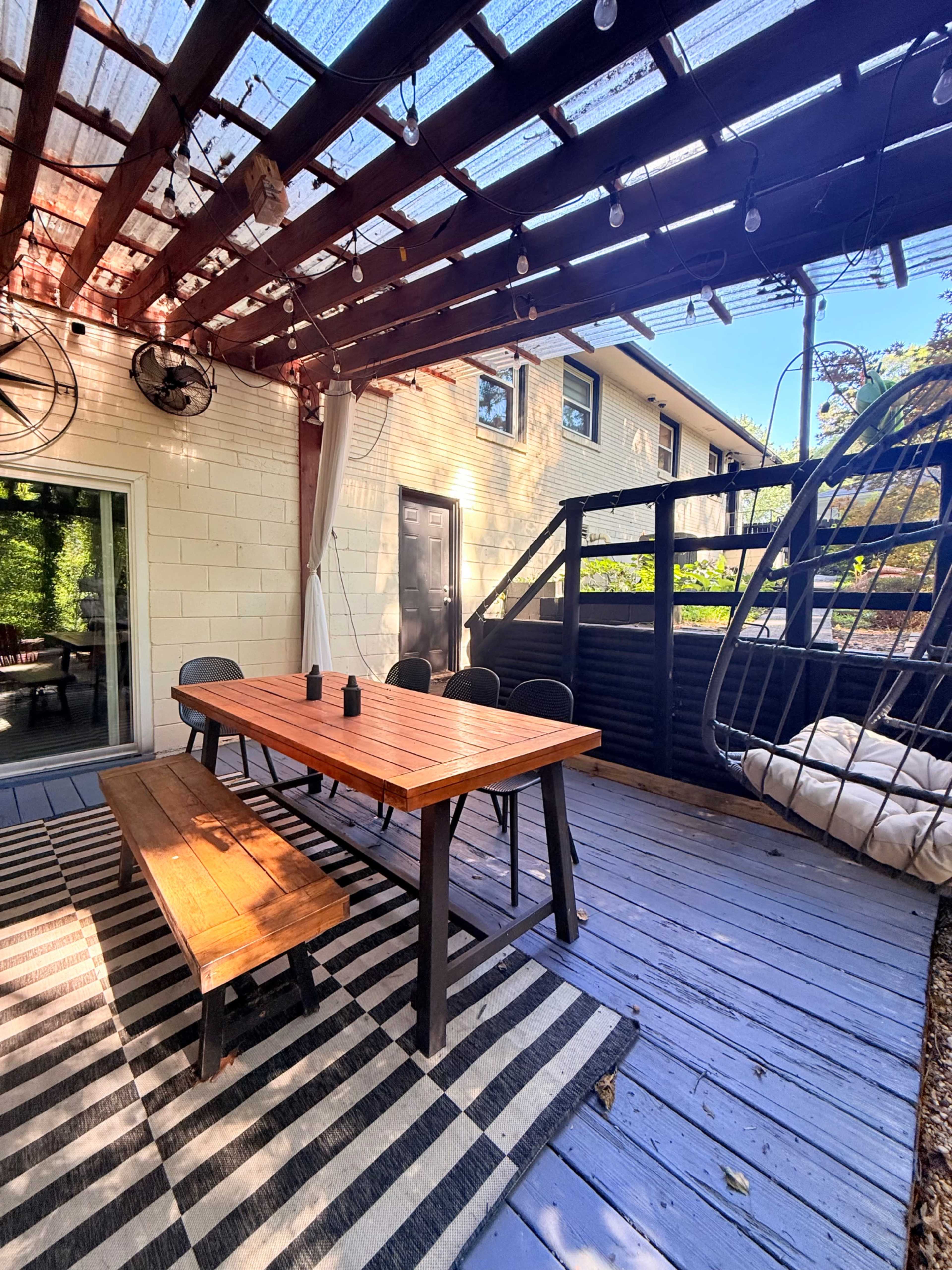 A wooden dining table with black chairs is set up under a covered patio, with a hanging chair and a patterned rug on a wooden deck.