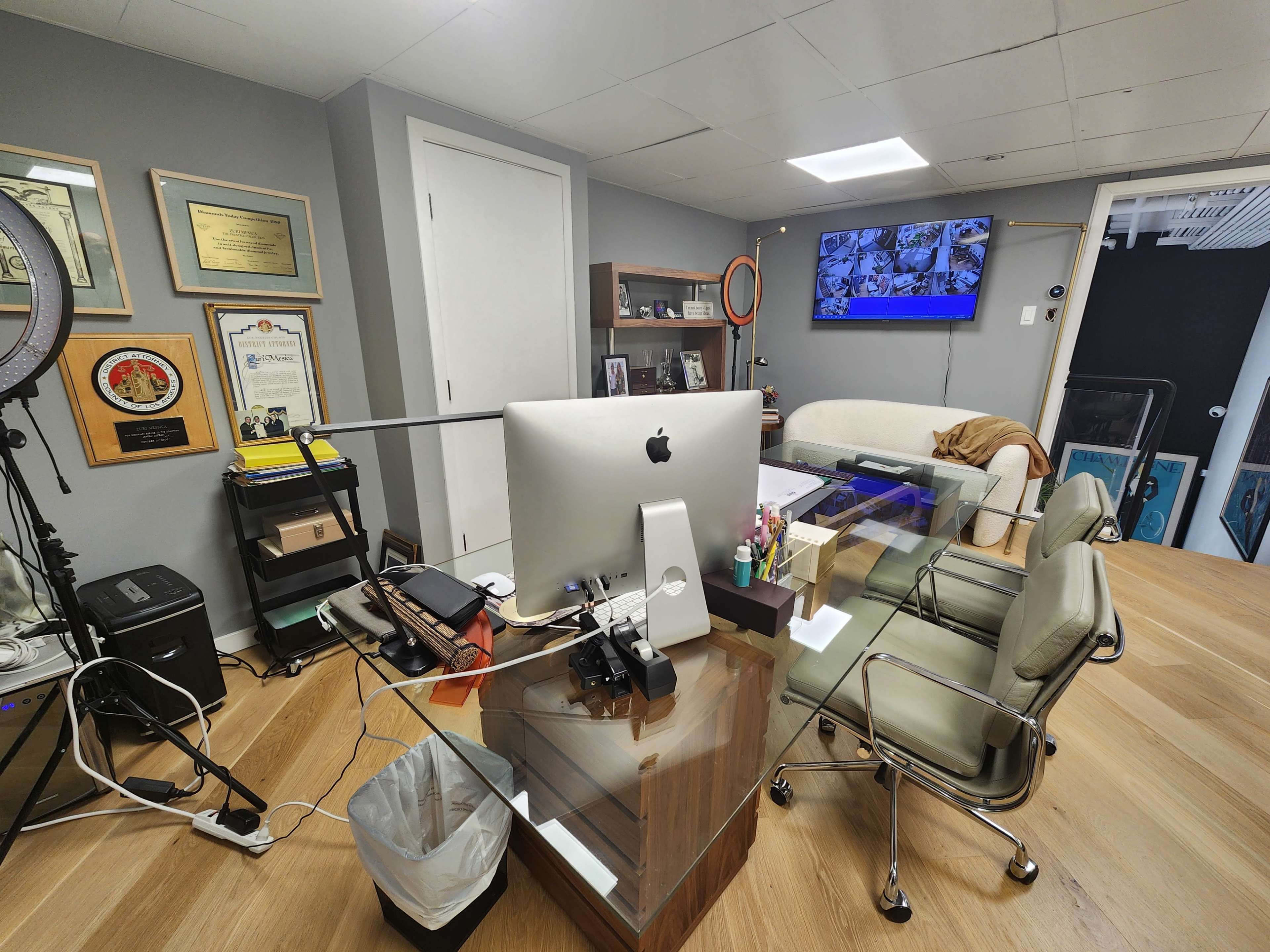 The image shows a modern office space featuring a large desk with an iMac, a comfortable chair, and various awards and a television mounted on the wall.