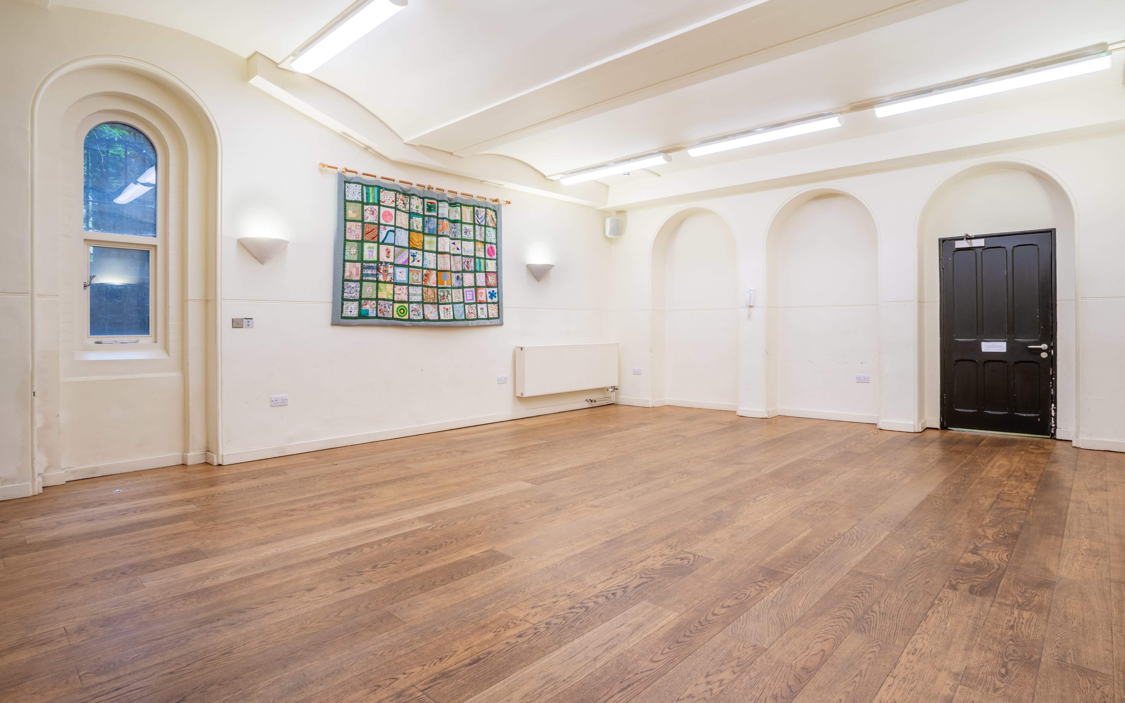 An empty room with wooden flooring, white walls, and a colorful mural displayed on one wall, along with a door on the right and two windows on the left.
