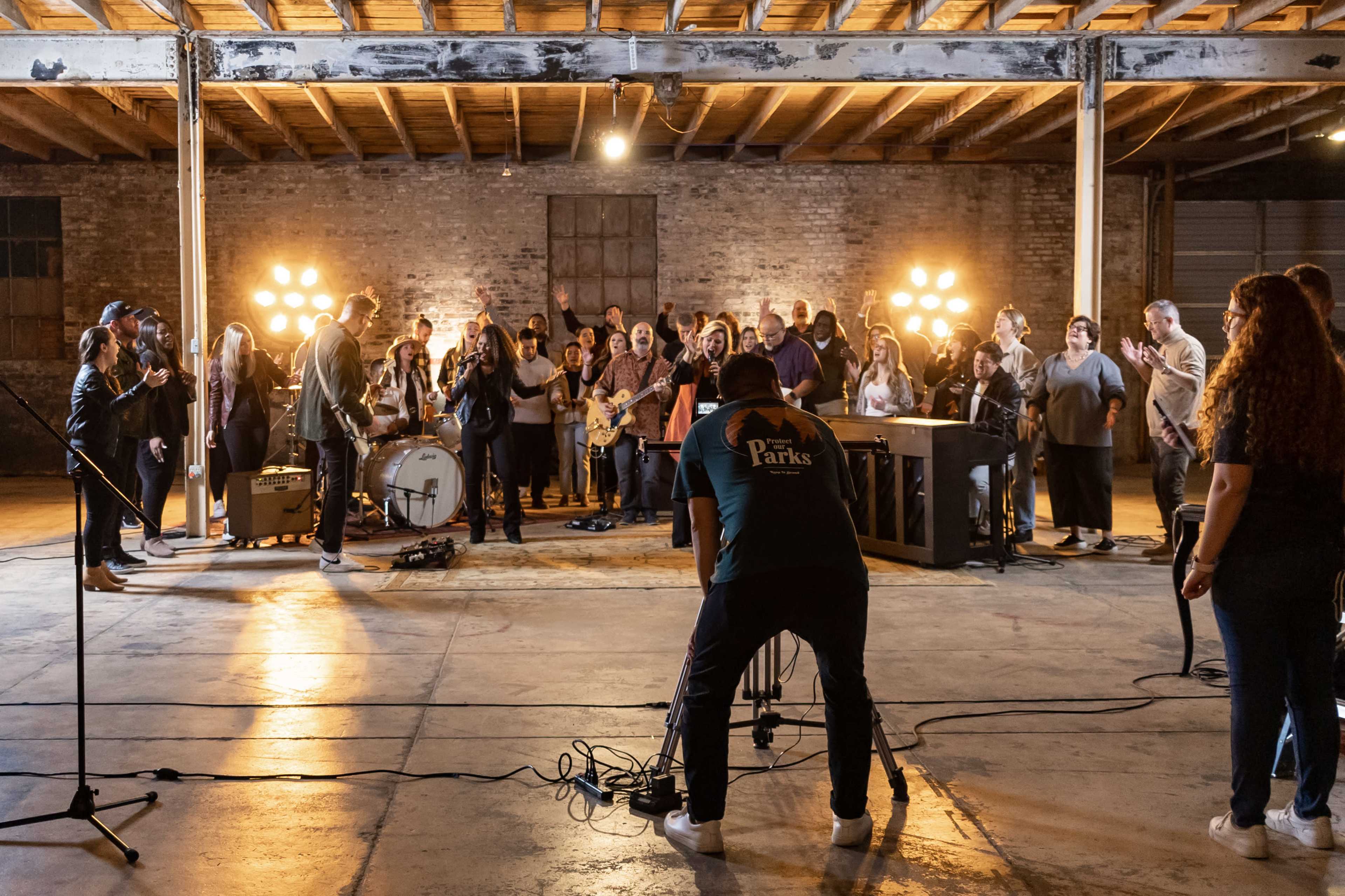 A large group of musicians and attendees are gathered in a spacious industrial room for a live music performance, with various instruments set up on stage and multiple bright lights illuminating the scene.