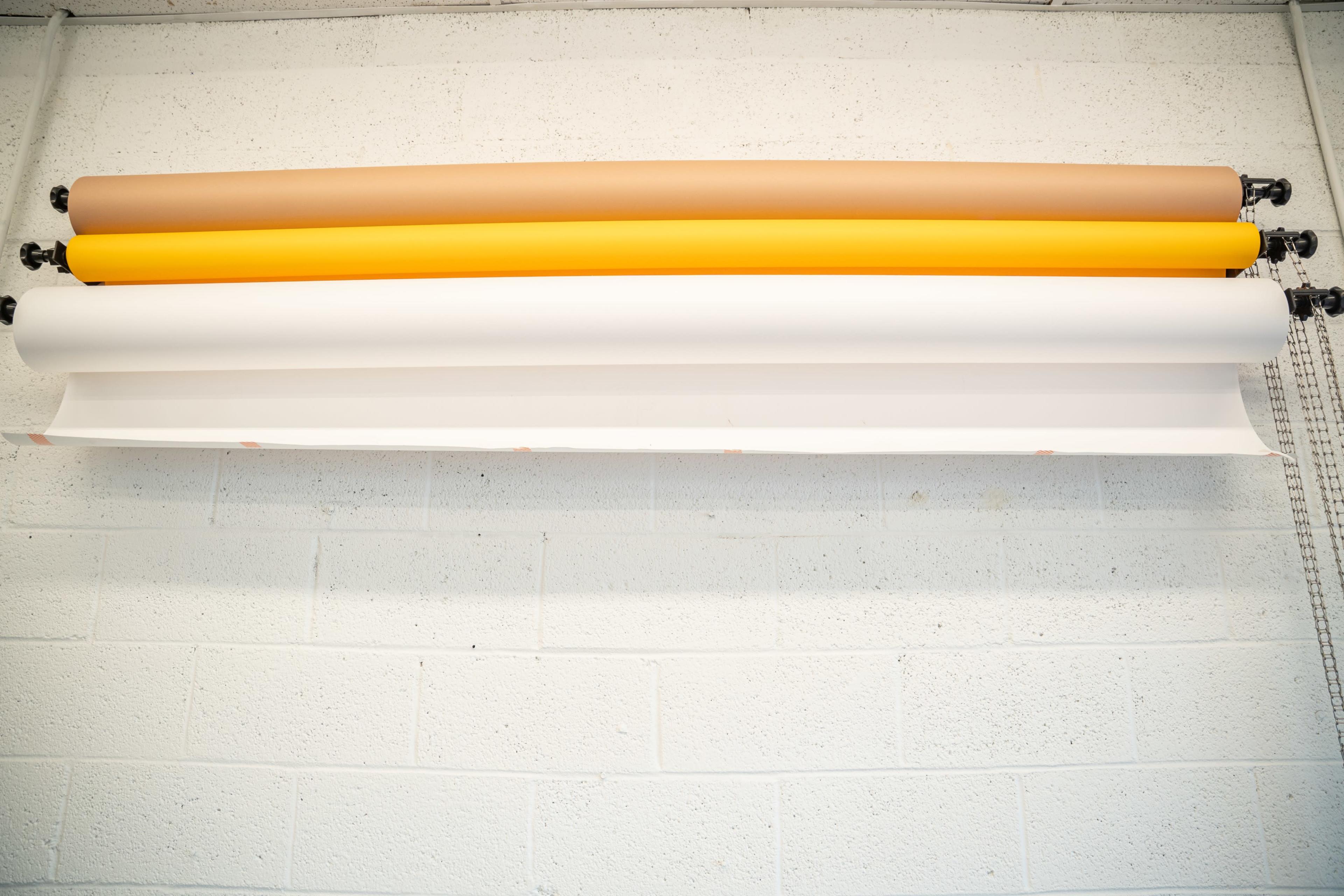 Several rolls of colored paper positioned horizontally on a wall-mounted rack against a textured white brick background.