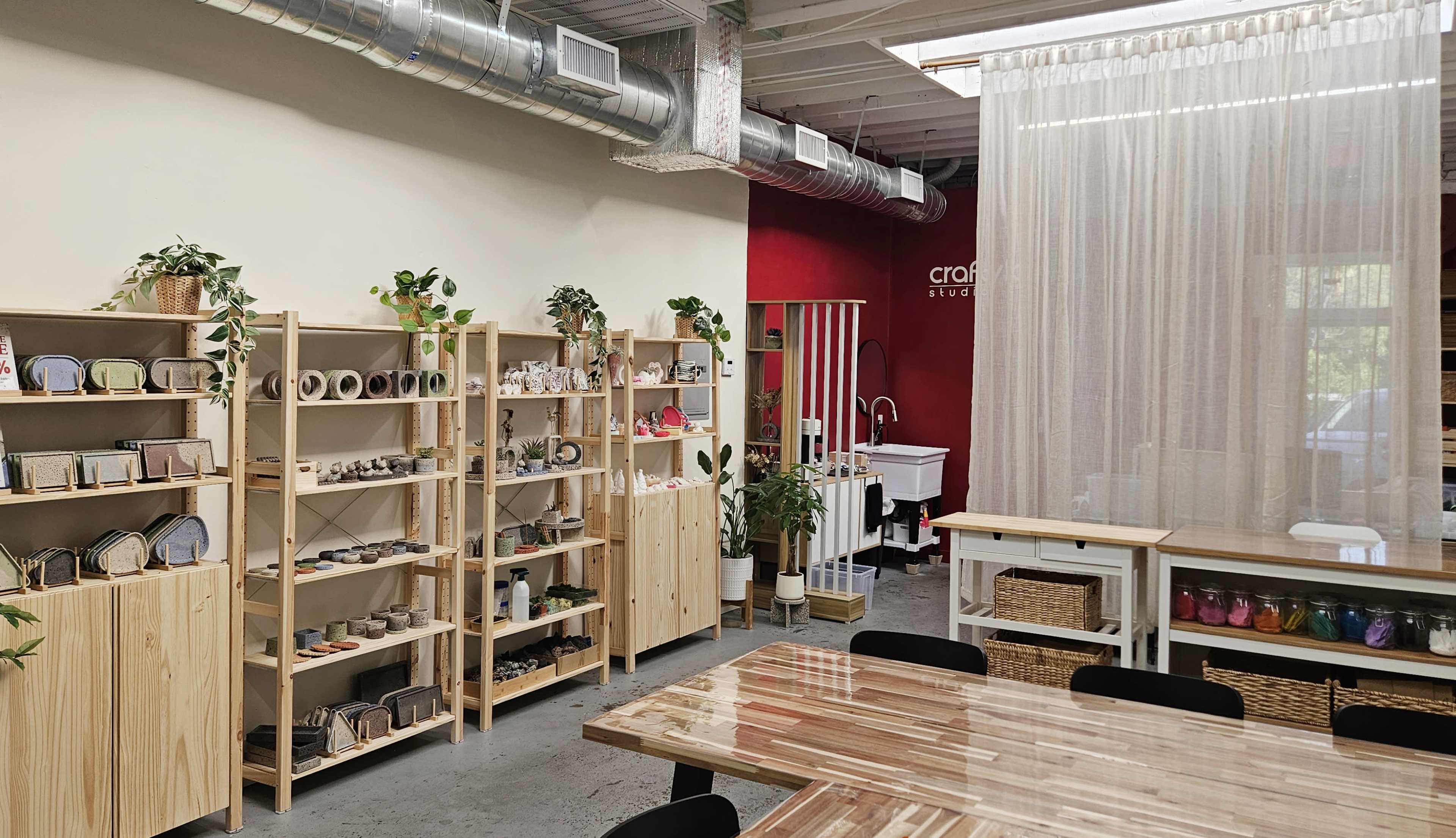 A bright, organized craft studio features wooden shelves filled with various handmade items, a large communal table, and bright red walls complemented by a curtained area.