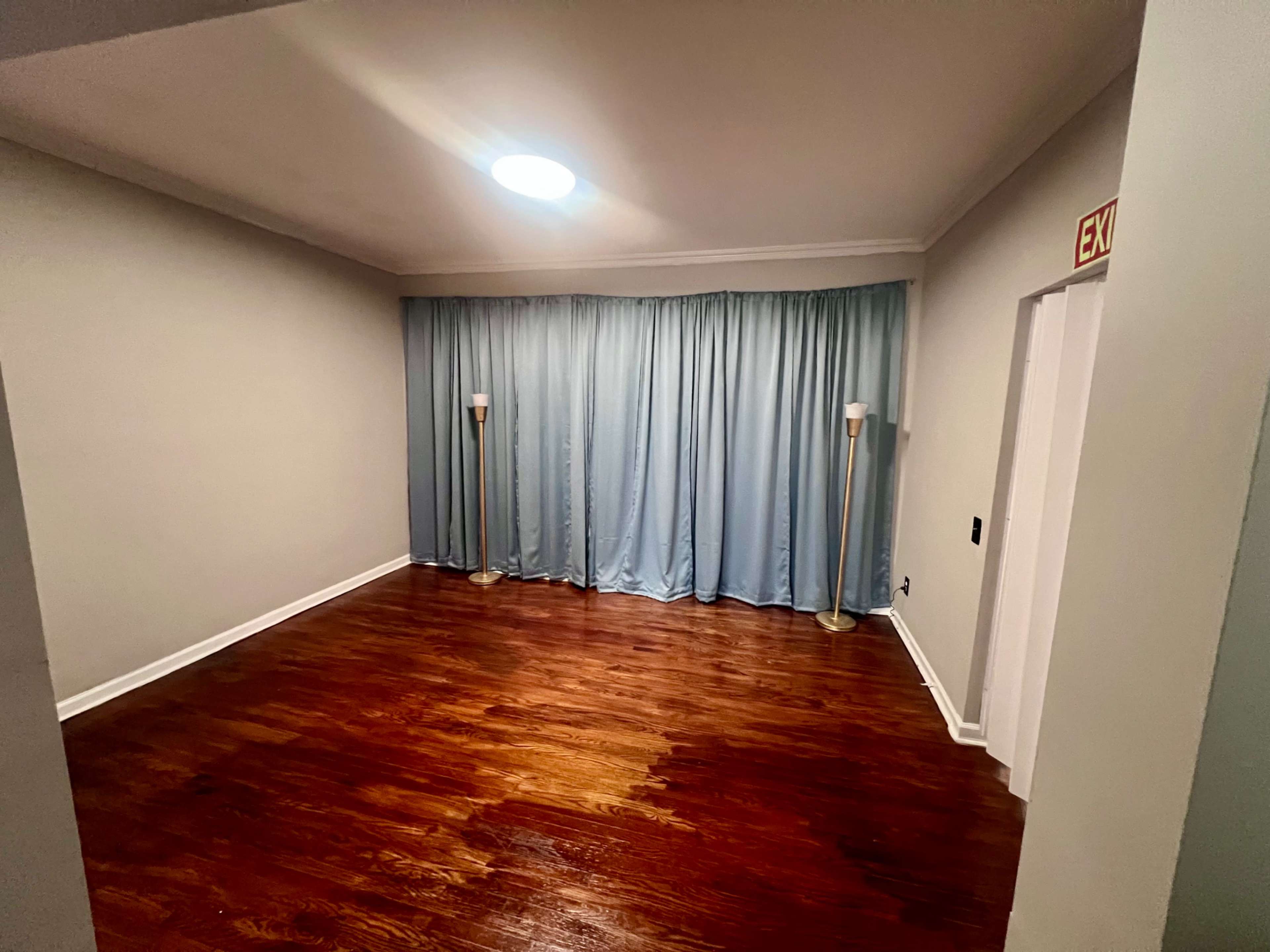 The image shows a sparsely furnished room with hardwood floors, light blue curtains, and two floor lamps near the curtains.