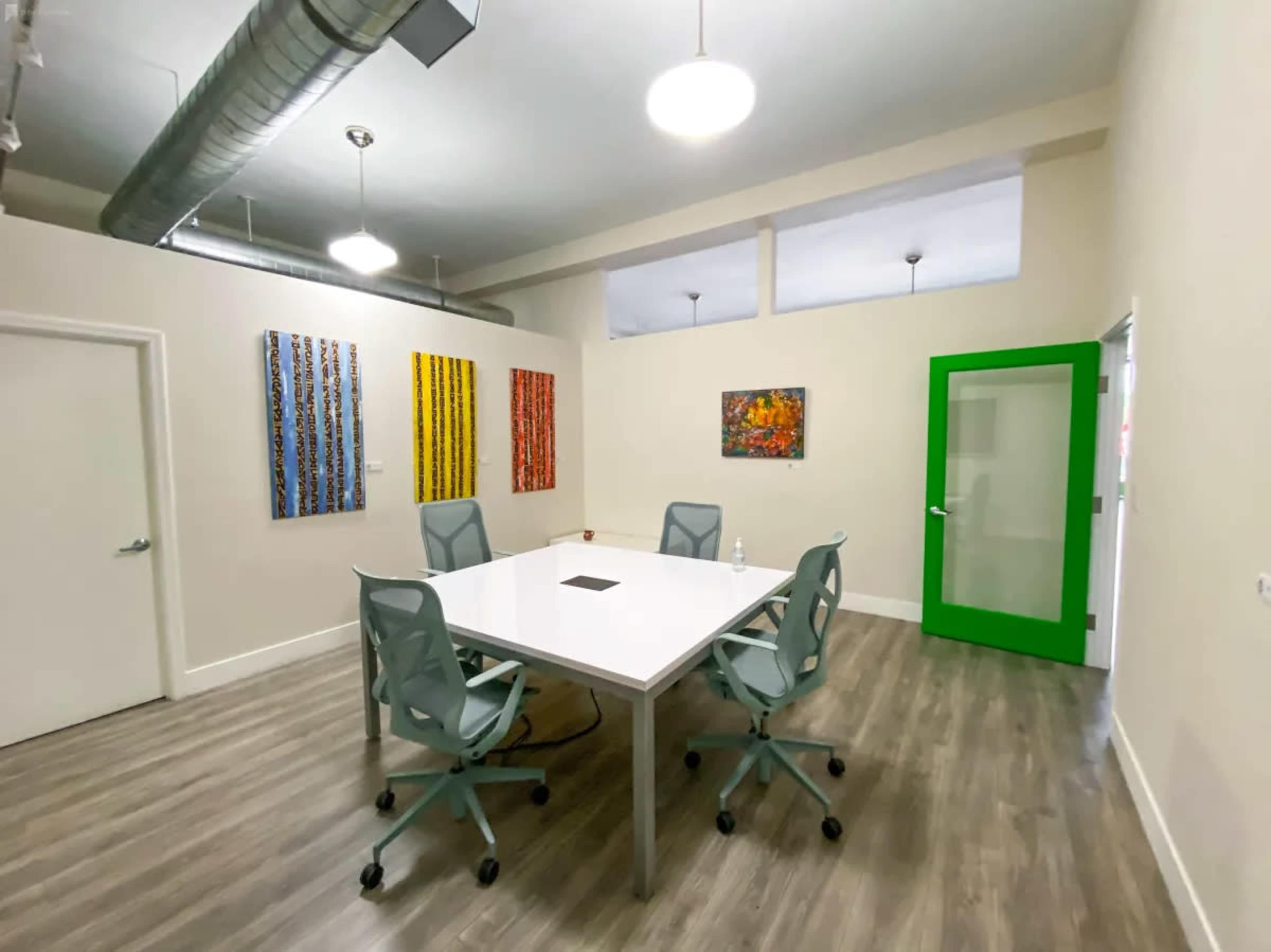 A modern meeting room with a white table, six gray chairs, and colorful paintings on the walls, featuring a green glass door.