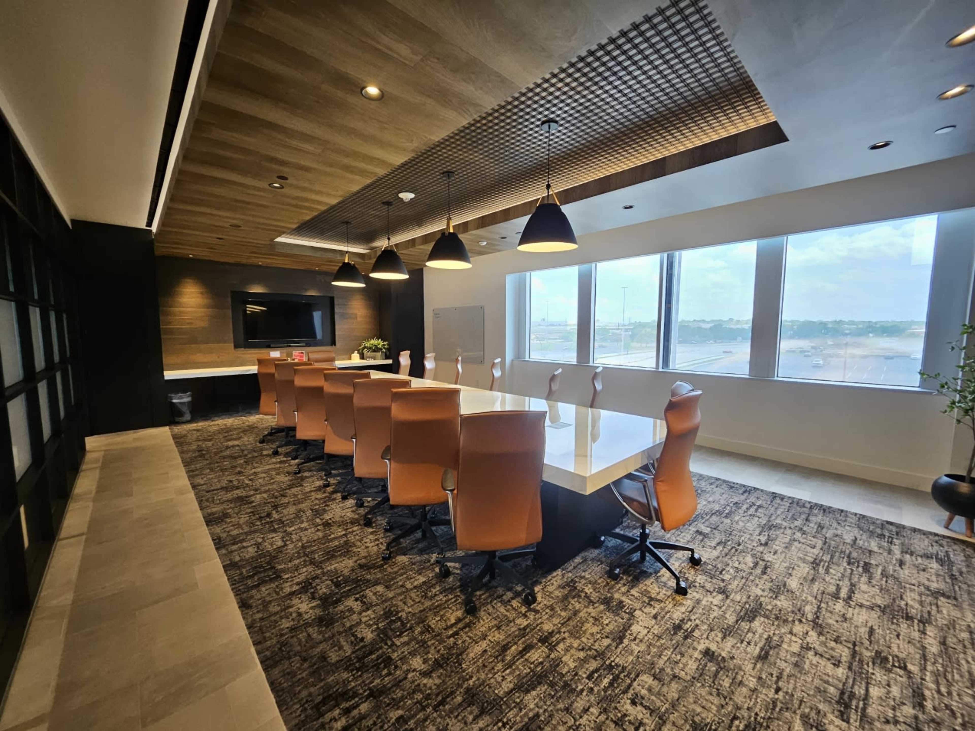 A modern conference room features a long white table with ergonomic orange chairs, large windows providing natural light, and a wooden ceiling design.