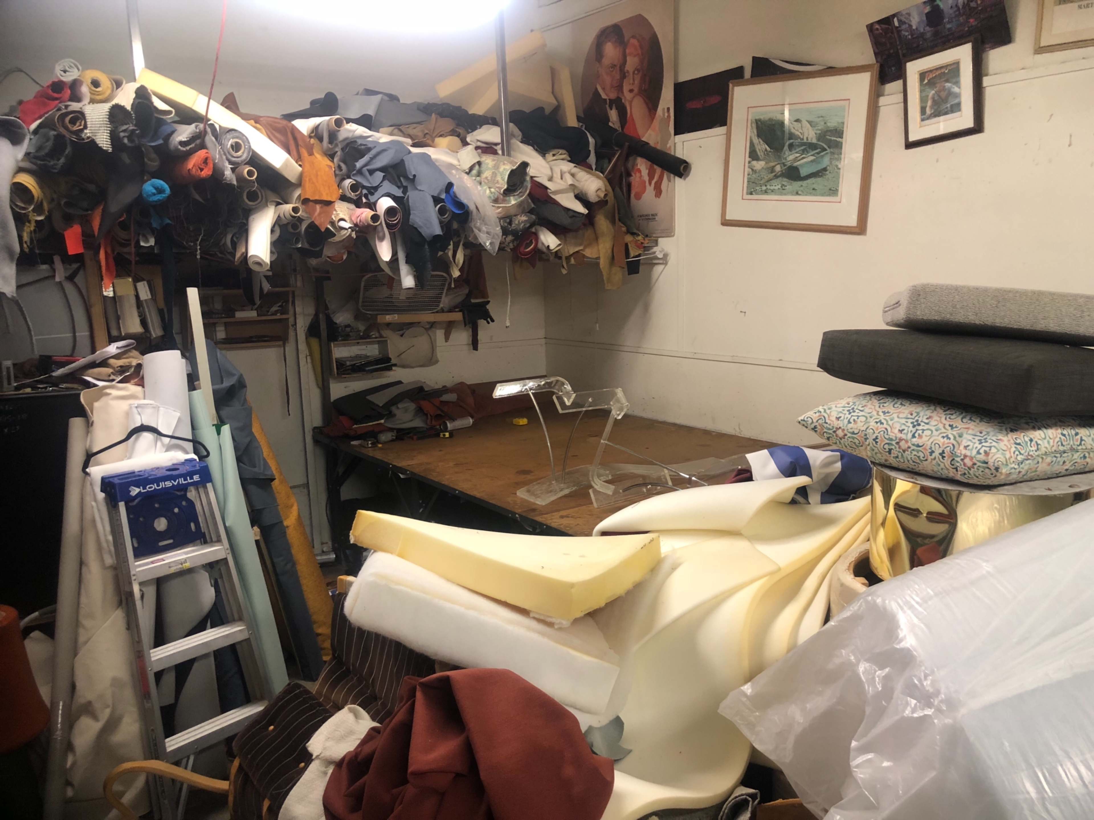 The image shows a cluttered workspace filled with rolls of fabric, stacks of cushions, and various tools, with a large table in the center.