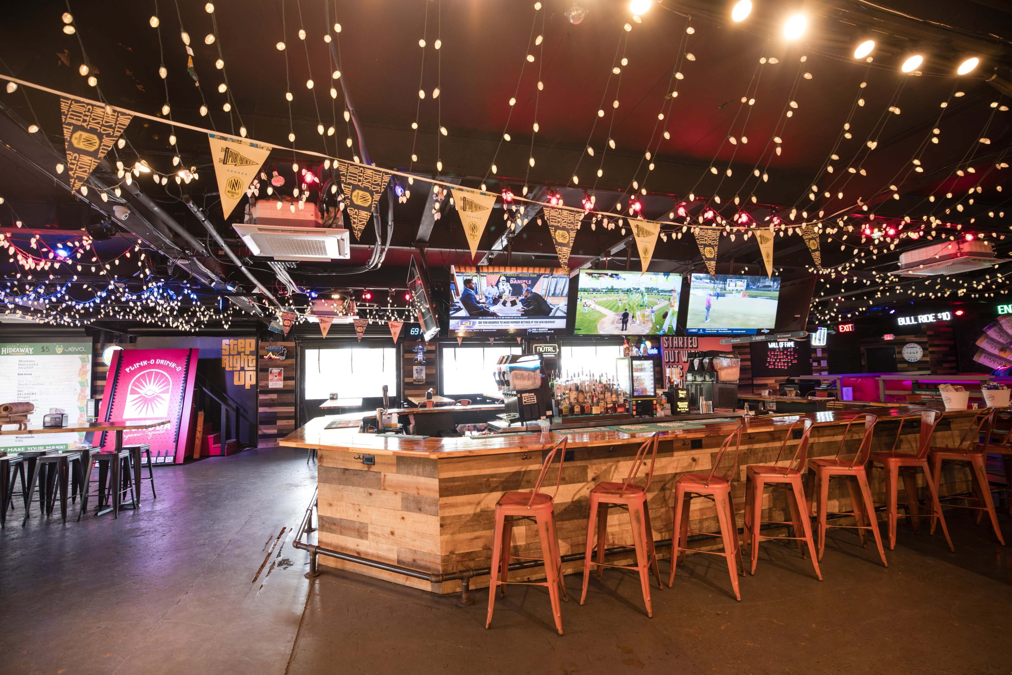 The image shows a lively bar area with wooden seating, illuminated by string lights and decorated with banners, featuring multiple screens displaying sports events.