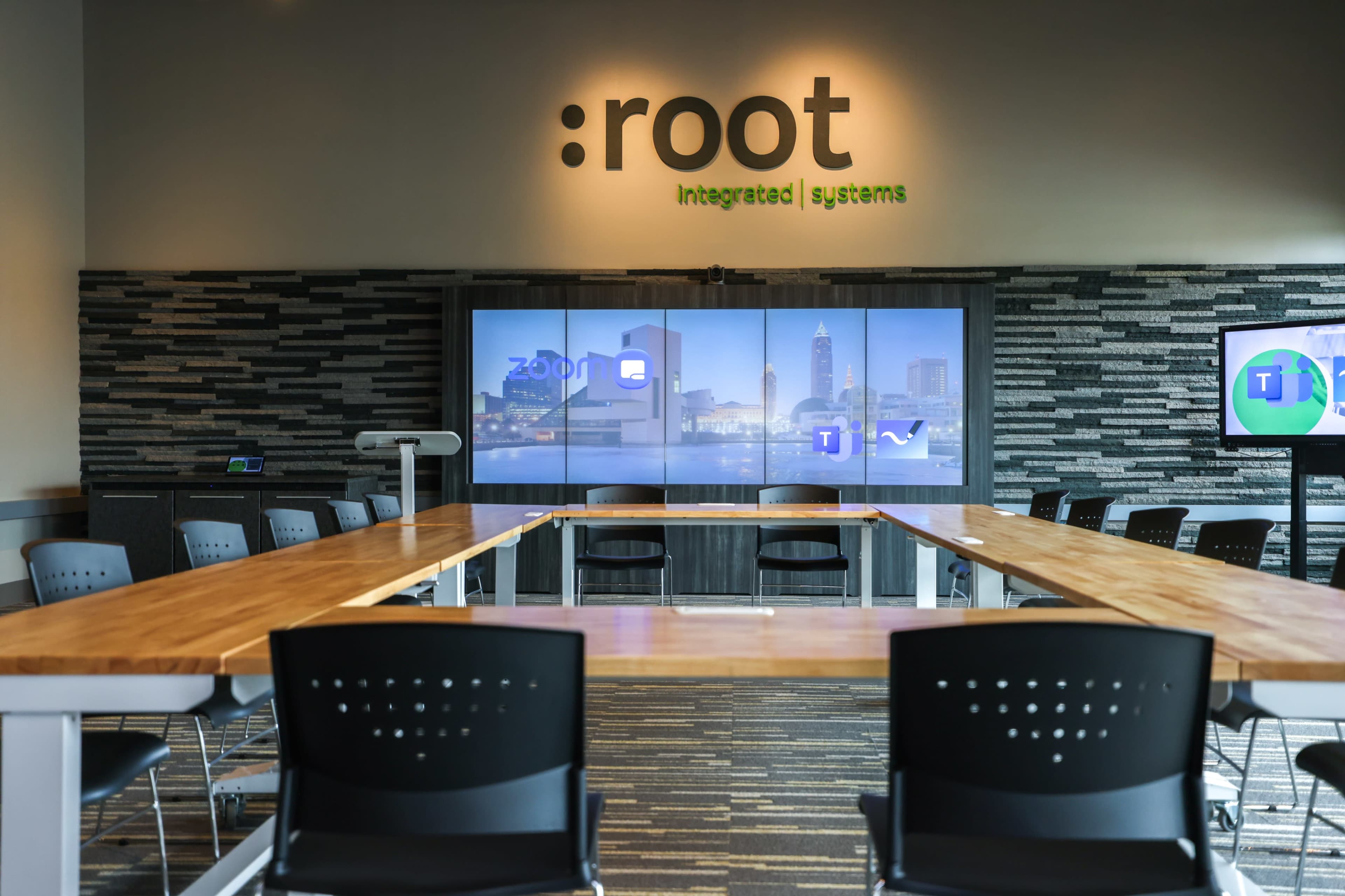 A modern conference room with a large wooden table, black chairs, and a screen displaying a city skyline and logos for Zoom and Microsoft Teams.
