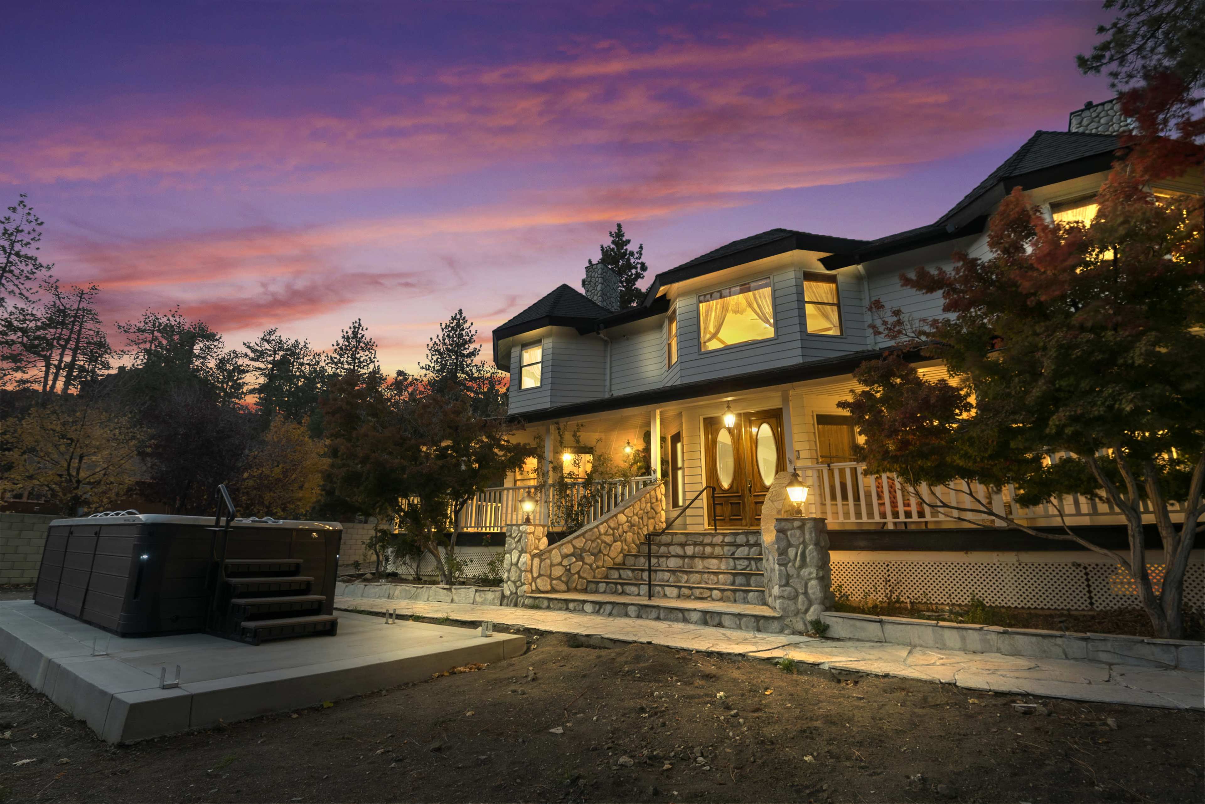 A large house with multiple stories is illuminated under a sunset sky, featuring a stone staircase and a hot tub on a concrete patio.