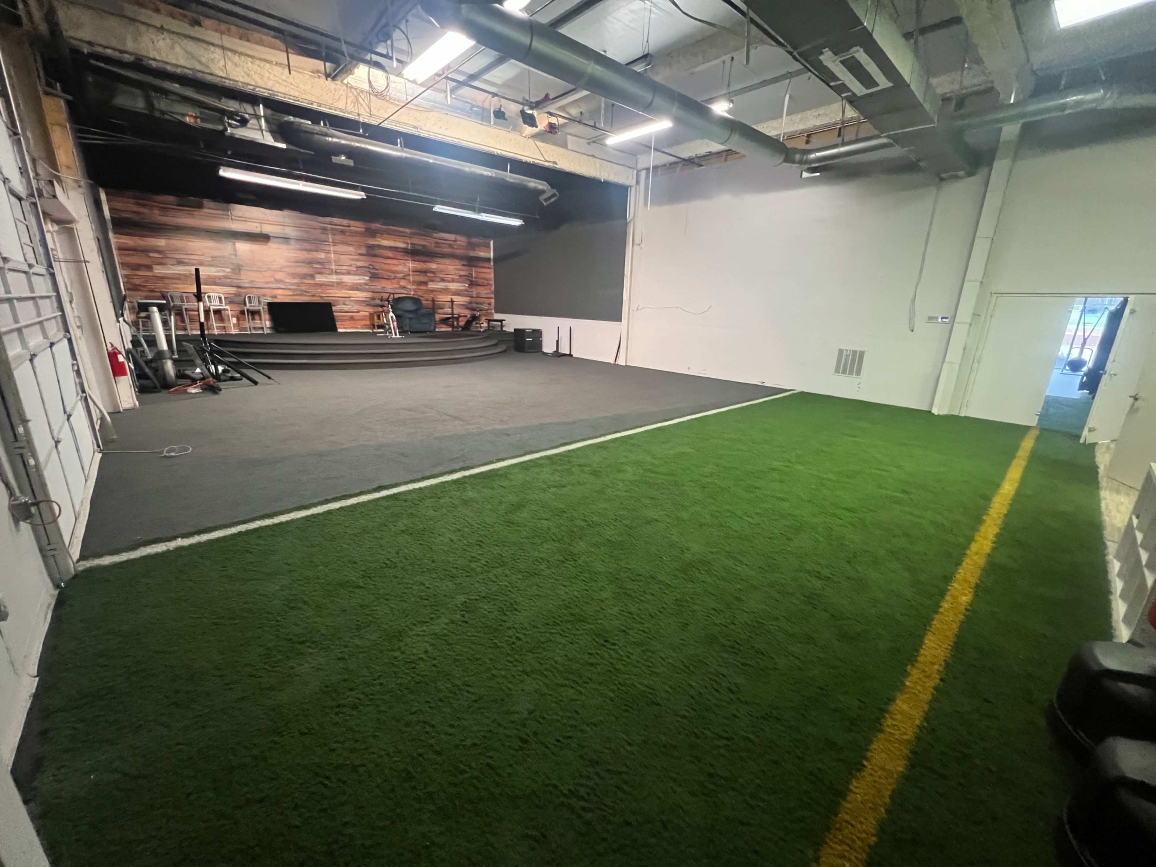 The image shows a spacious indoor training facility with a section of artificial turf and a wooden-paneled wall in the background.