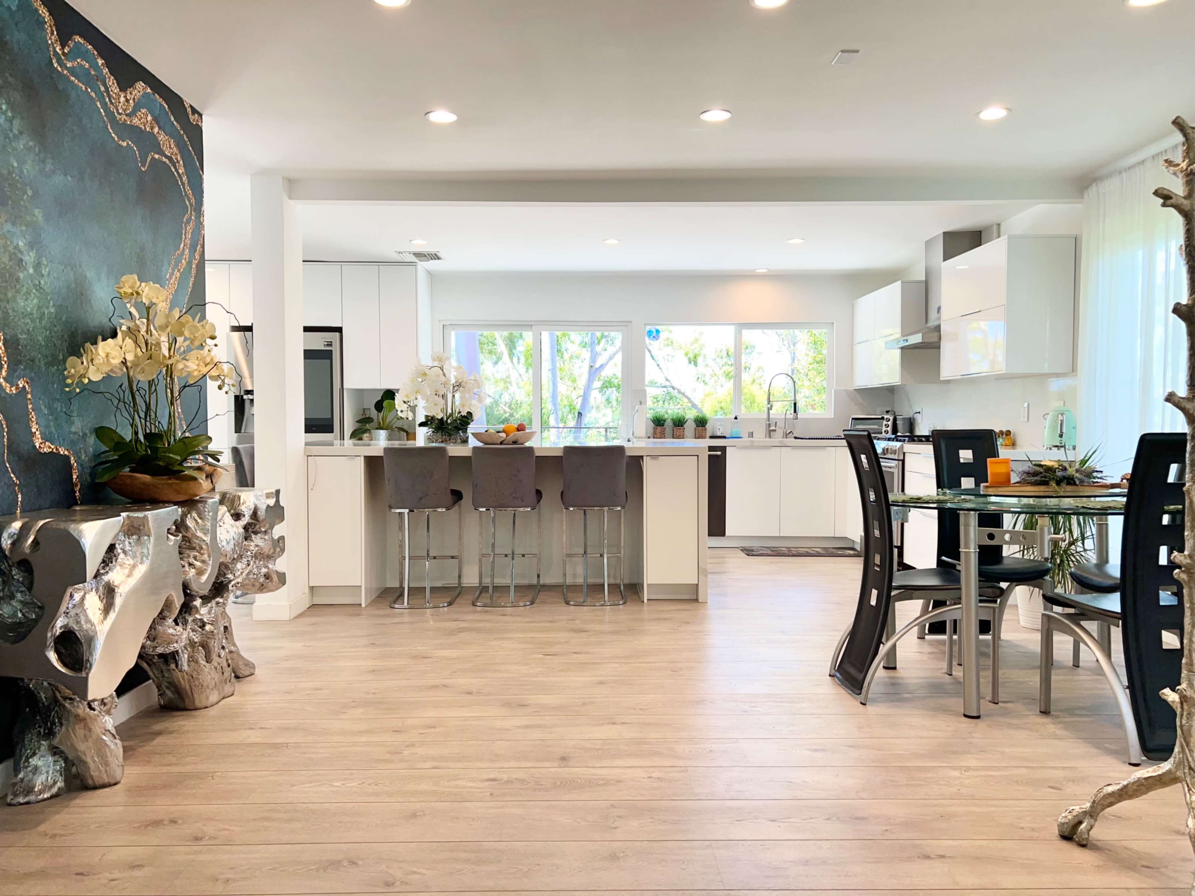 The image shows a modern kitchen and dining area with white cabinetry, a large window allowing natural light, and decorative elements including plants and a textured wall.