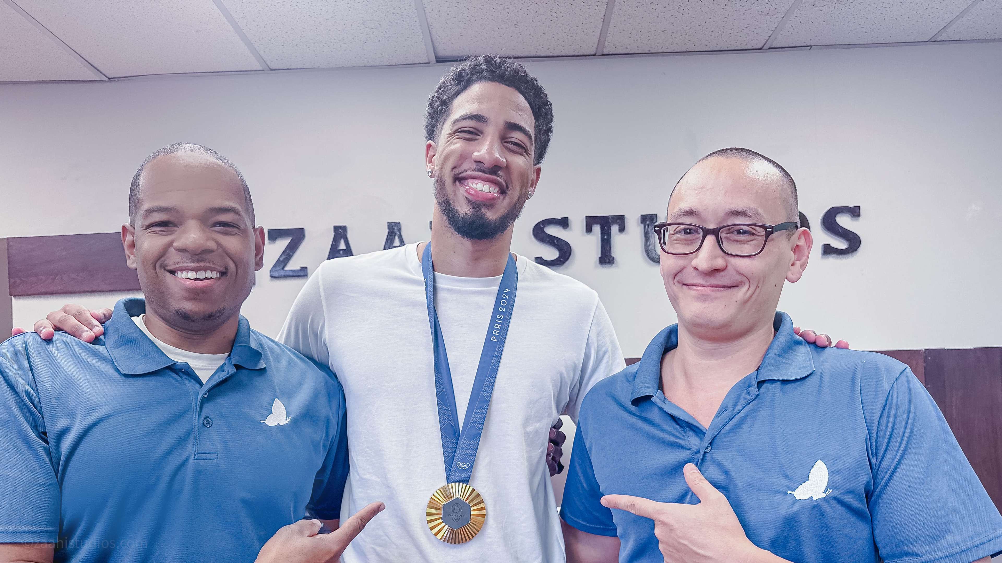Three men are posing together, with one man in the center wearing a medal, while the others are smiling and wearing matching blue shirts.