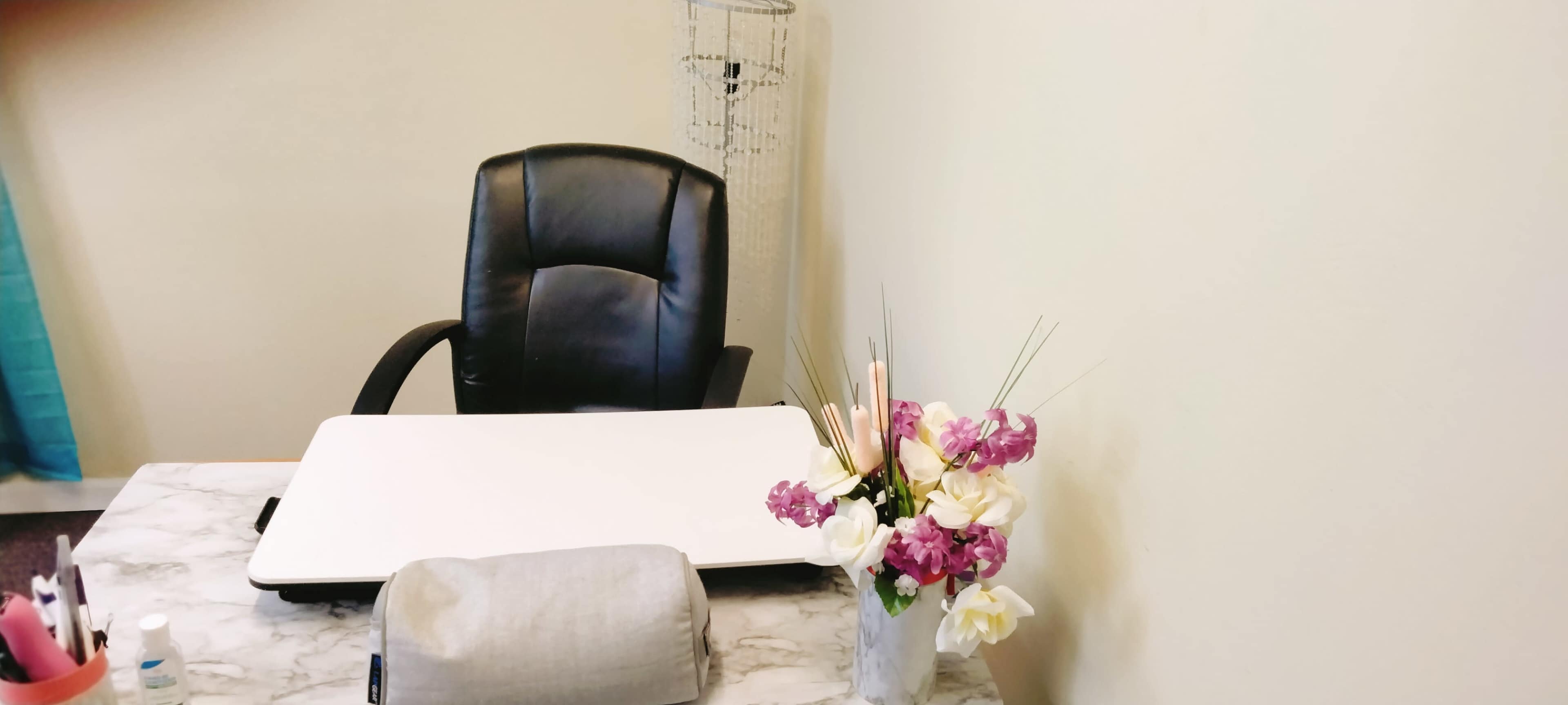 A black office chair is positioned next to a white desk adorned with a vase of flowers, a gray cushion, and various stationery items.
