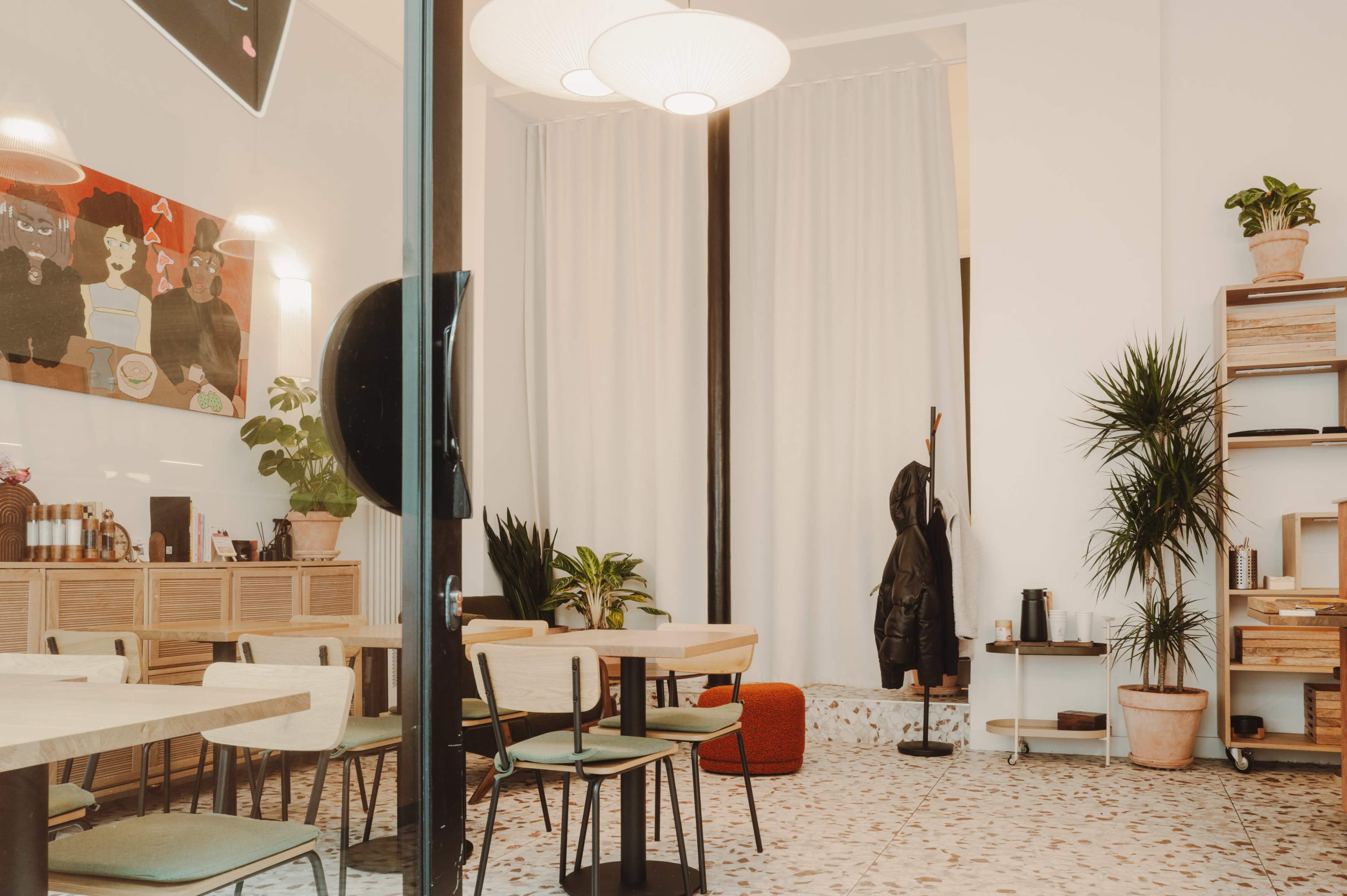 The interior of a modern café features wooden tables and chairs, decorative plants, and artwork on the wall, with bright light flooding in through large windows.