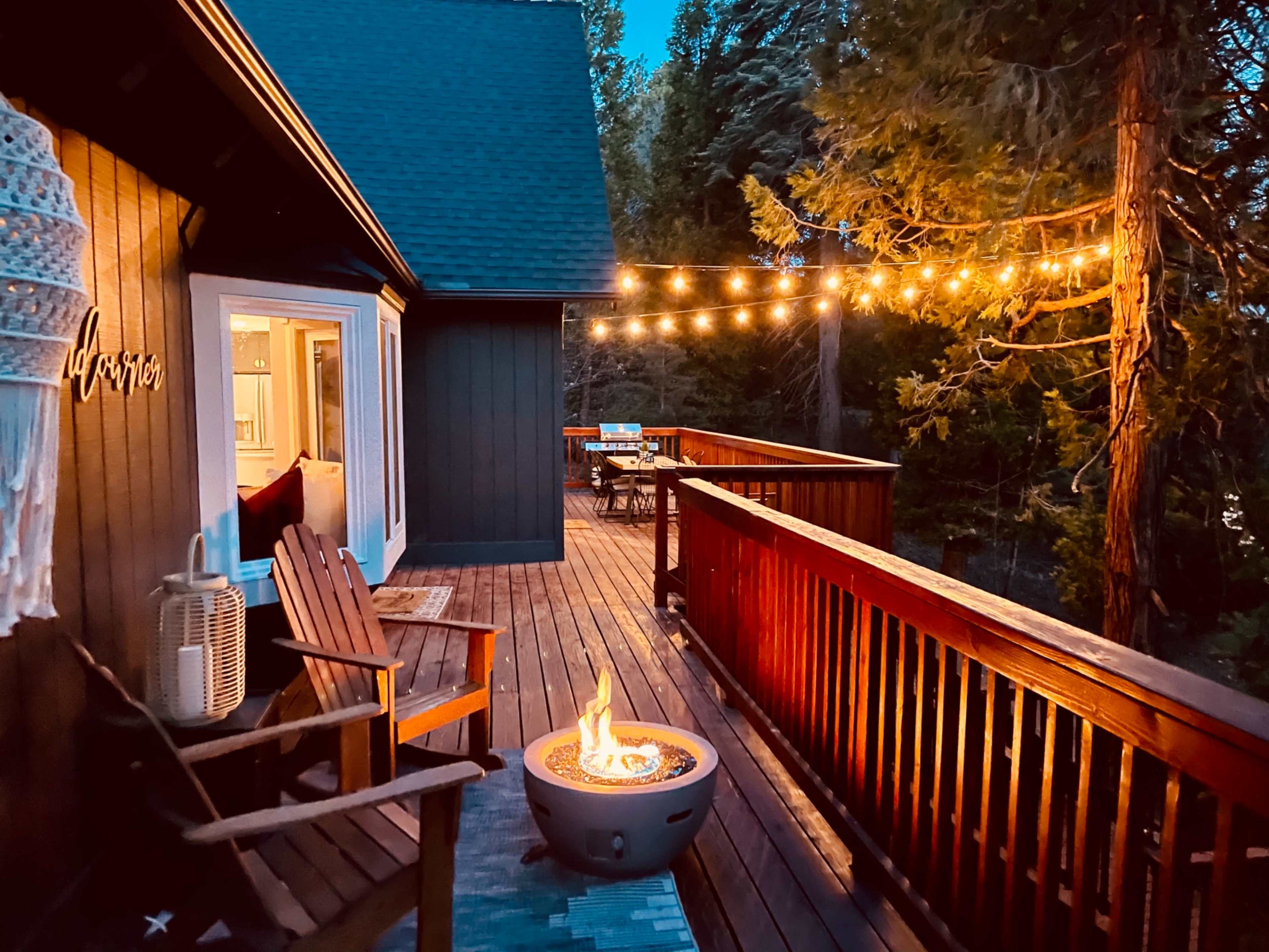 A wooden deck features a fire pit surrounded by chairs, string lights overhead, and a view of trees in the evening.