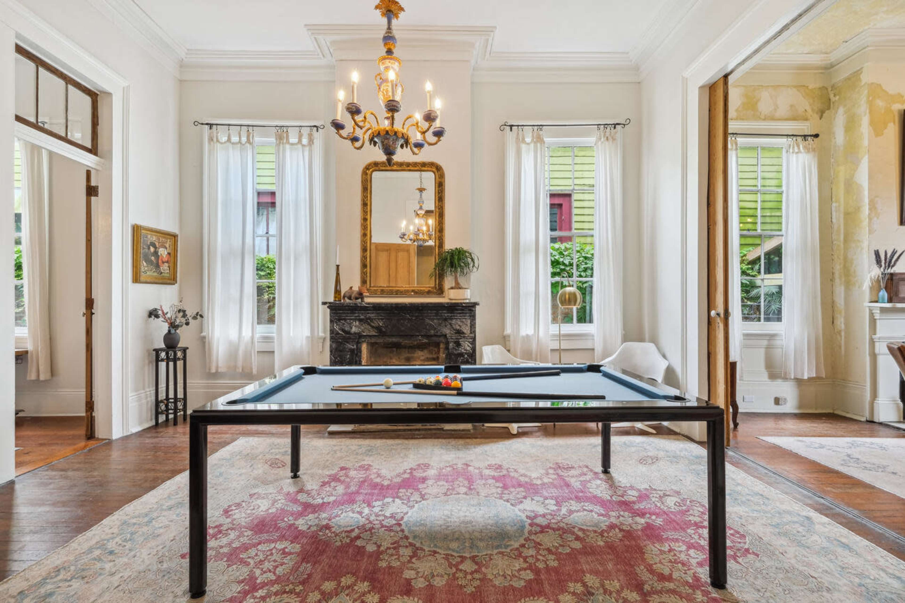 A billiard table is centered in a spacious room featuring a traditional chandelier, a large mirror, and tall windows that allow natural light to enter.