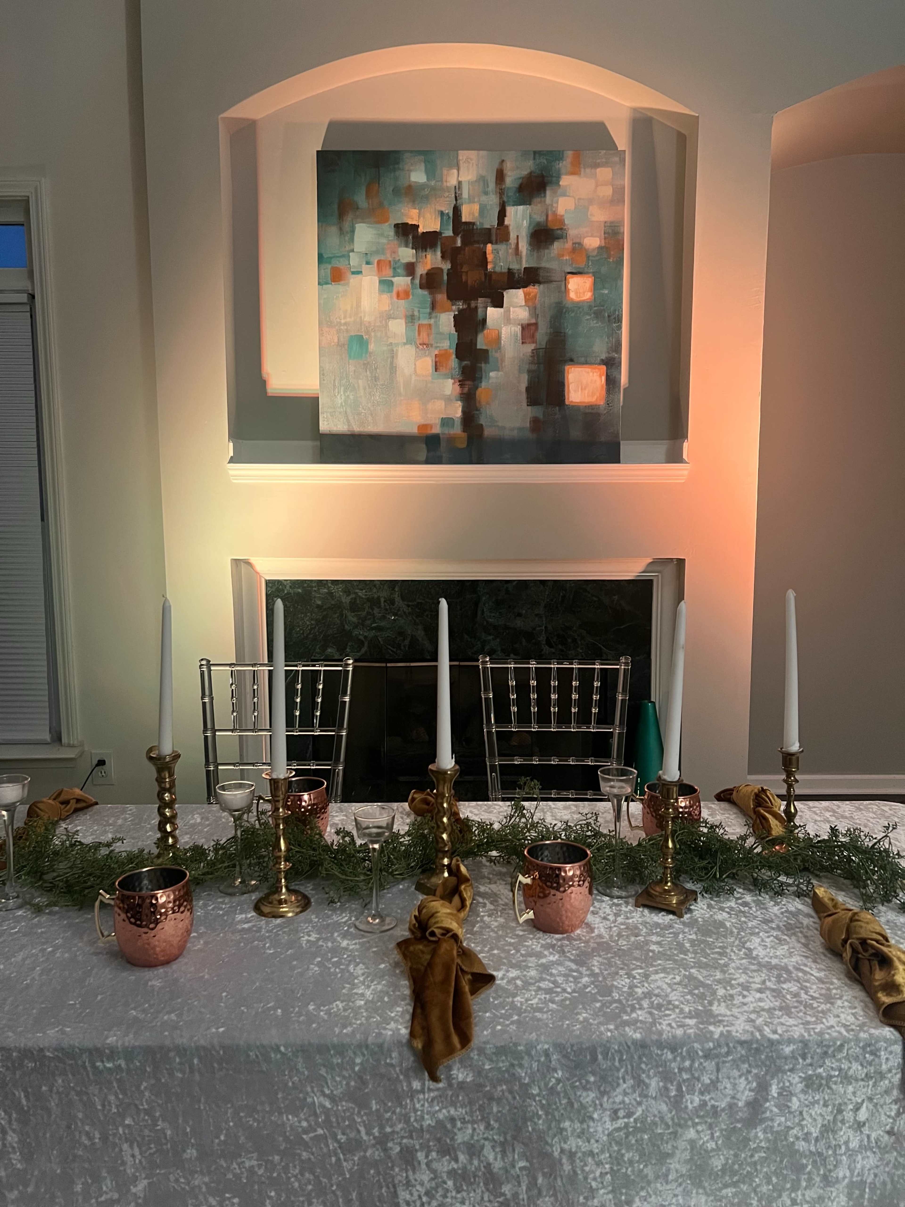 A dining table is set with gold and copper decor, white candles, and greenery in front of a modern abstract painting and a lit fireplace.