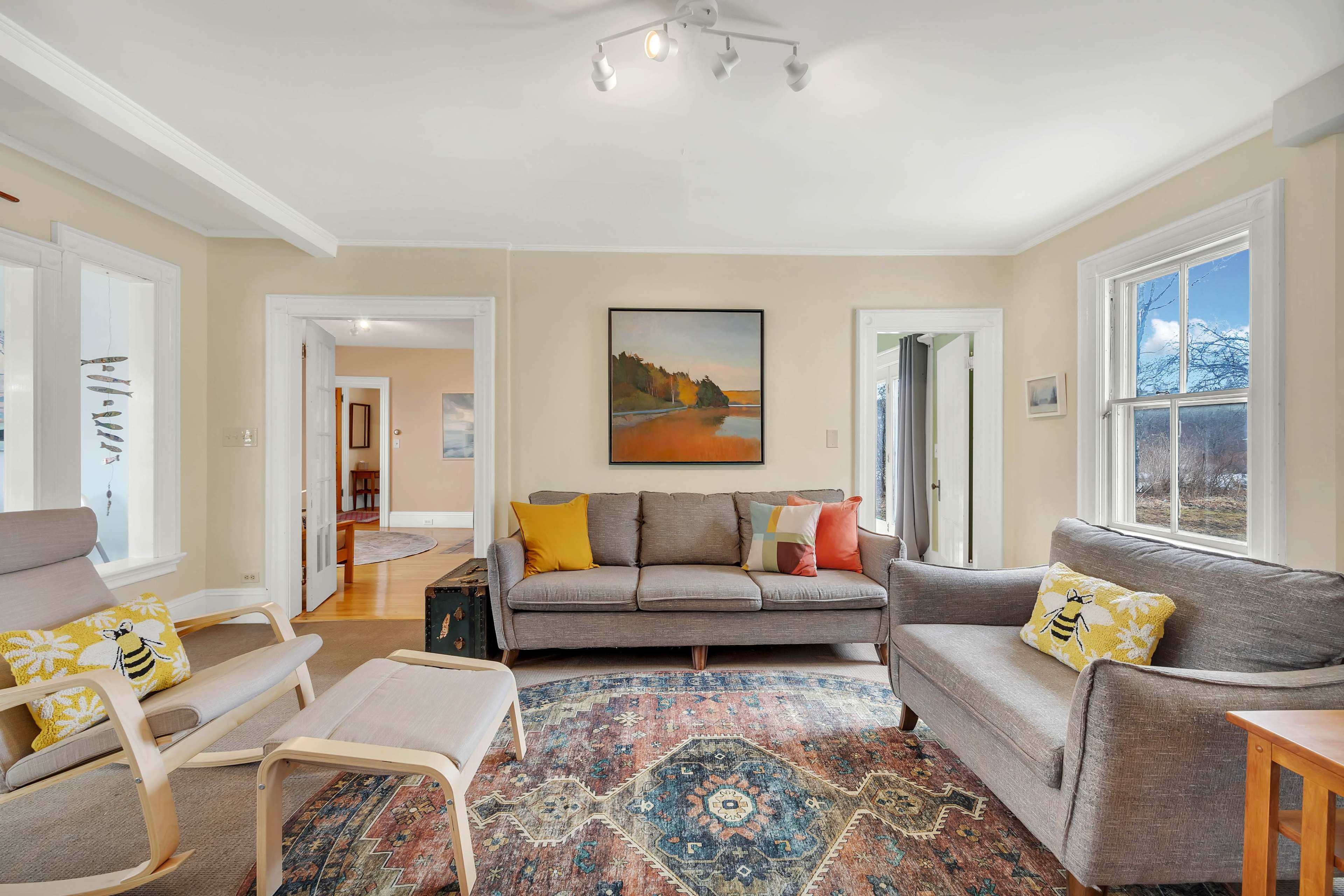 A light-filled living room features a gray sofa, a wooden chair with patterned pillows, and a colorful area rug, with large windows providing views of the outside.