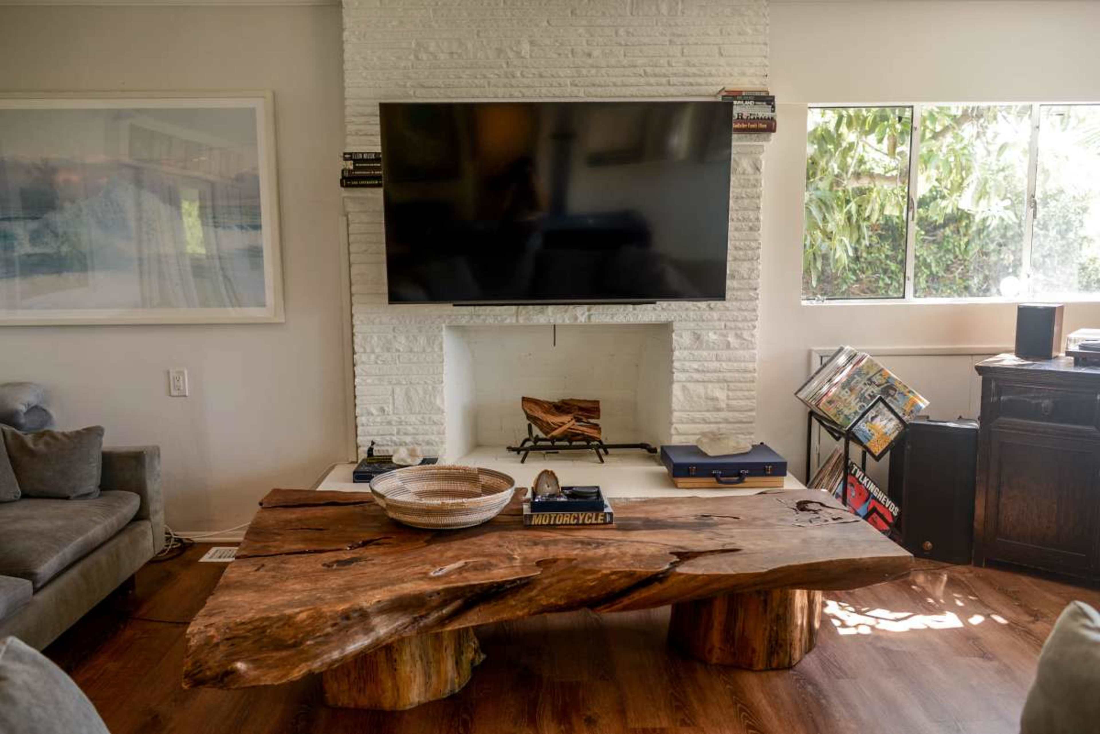 The image shows a modern living room with a large wooden coffee table, a flat-screen TV mounted above a white-bricked fireplace, and large windows letting in natural light.