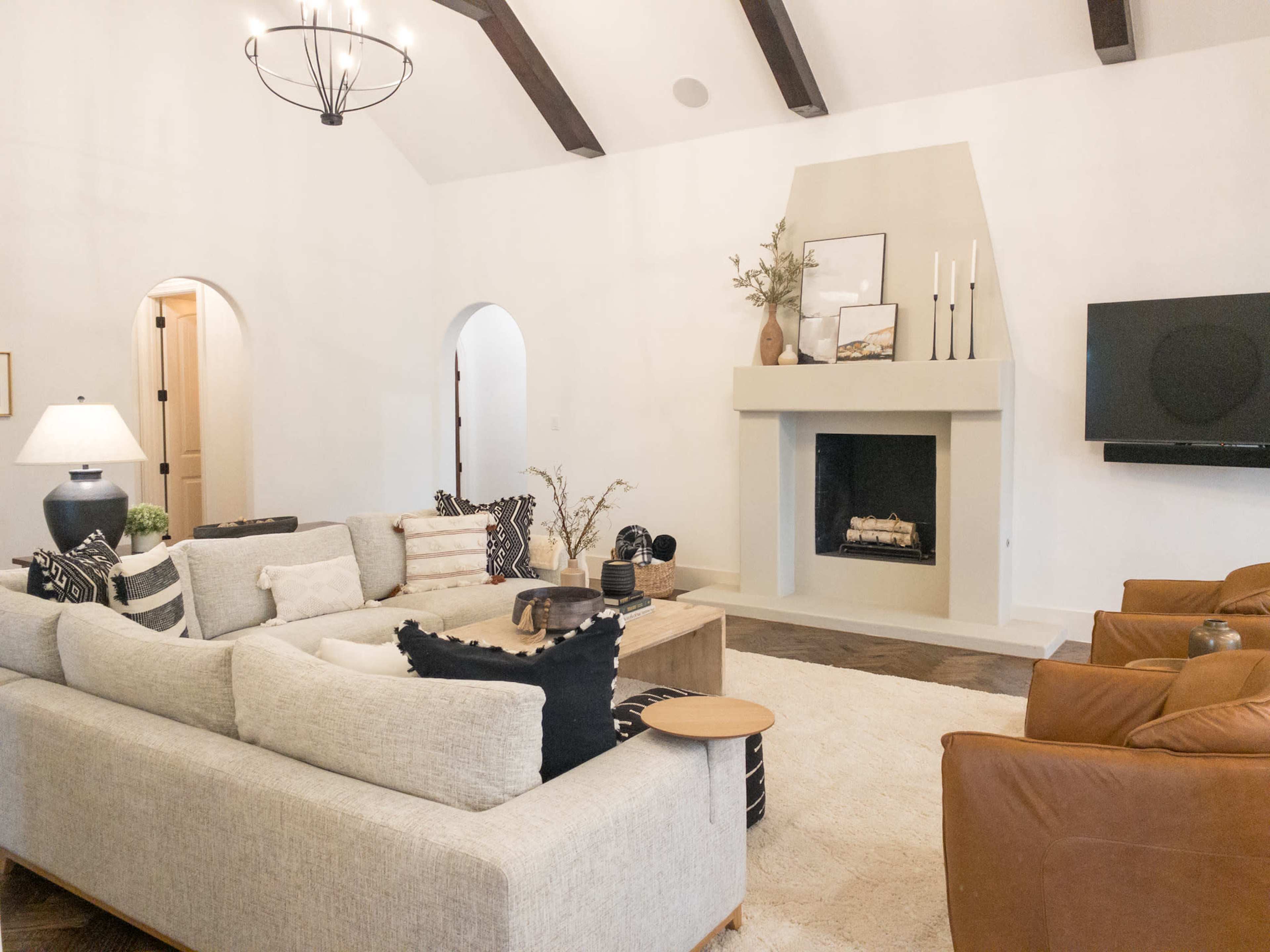 The living room features a large sectional sofa, a coffee table, and a fireplace with a minimalist design, set against a backdrop of white walls and wooden beams.
