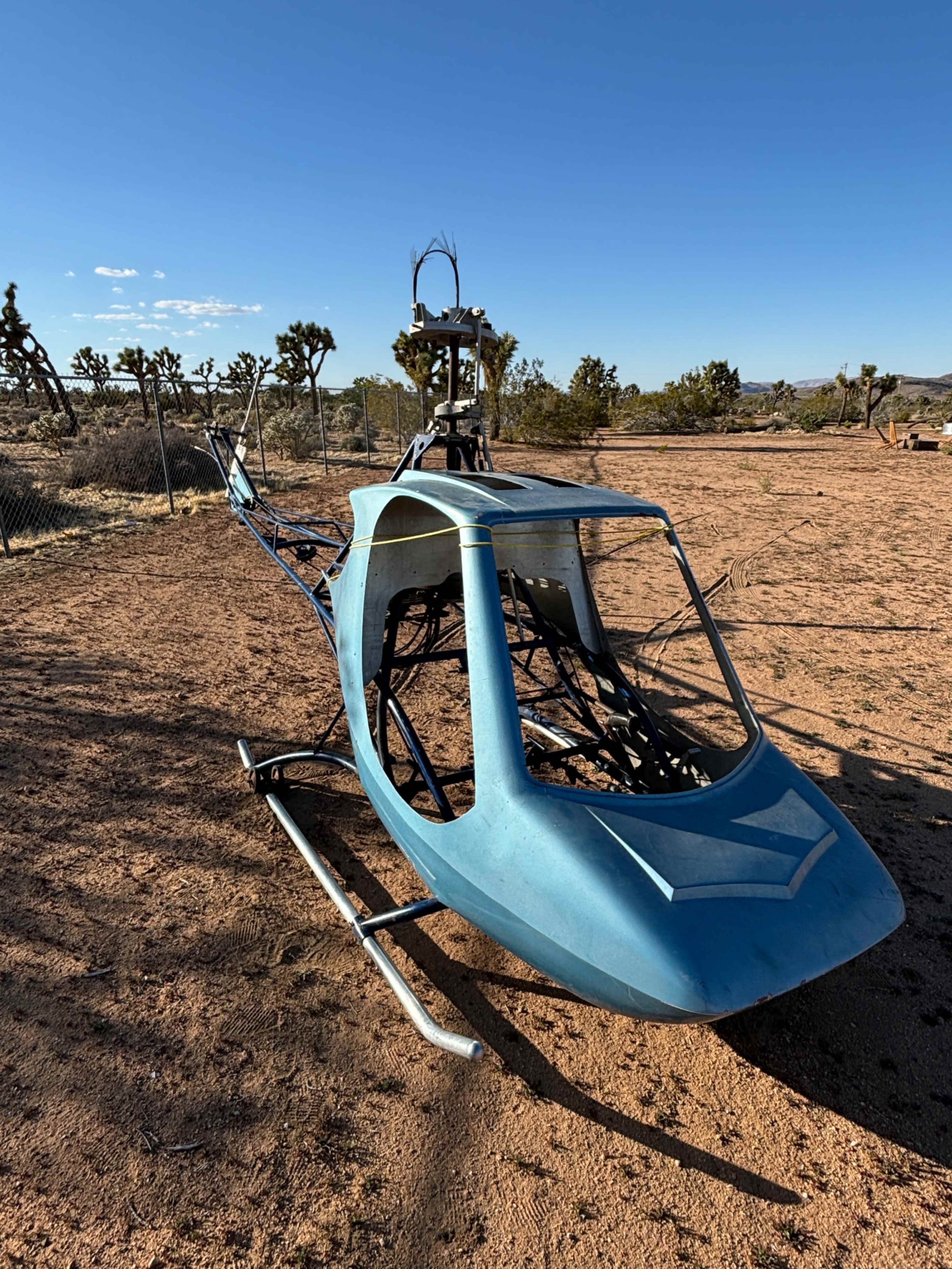 A blue helicopter fuselage is positioned on the ground in a desert landscape with sparse vegetation and clear blue skies.