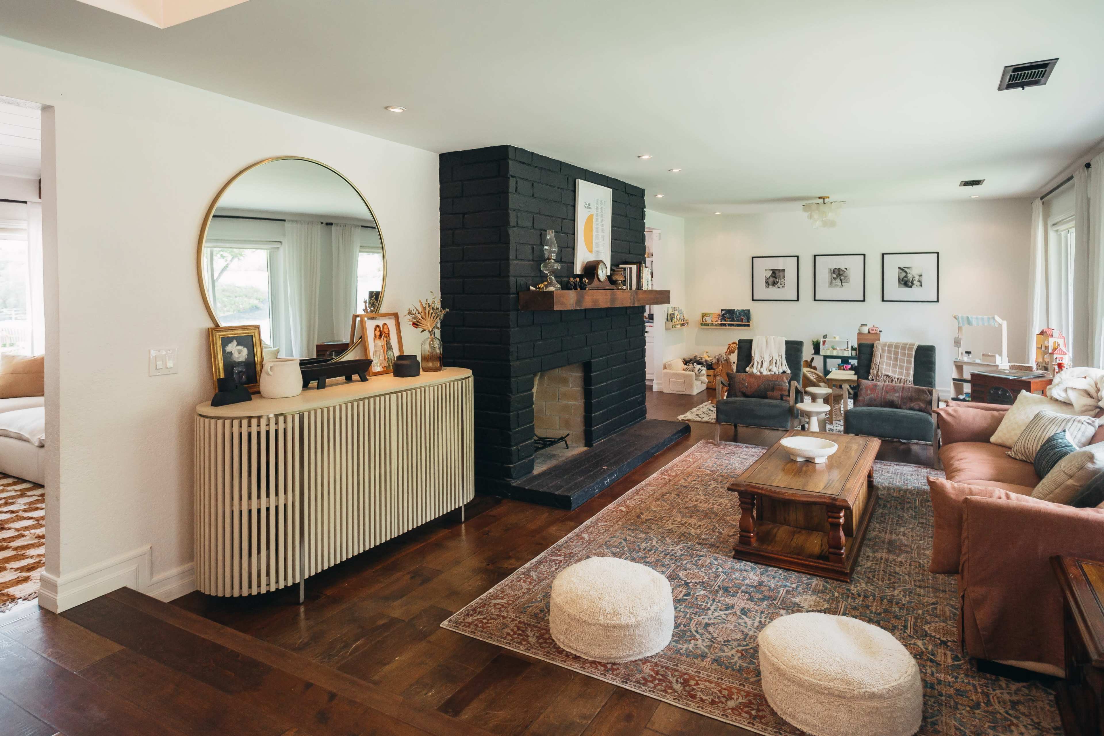 The image shows a living room with a black brick fireplace, a round mirror above a wooden console, and a mix of seating and decorative elements.