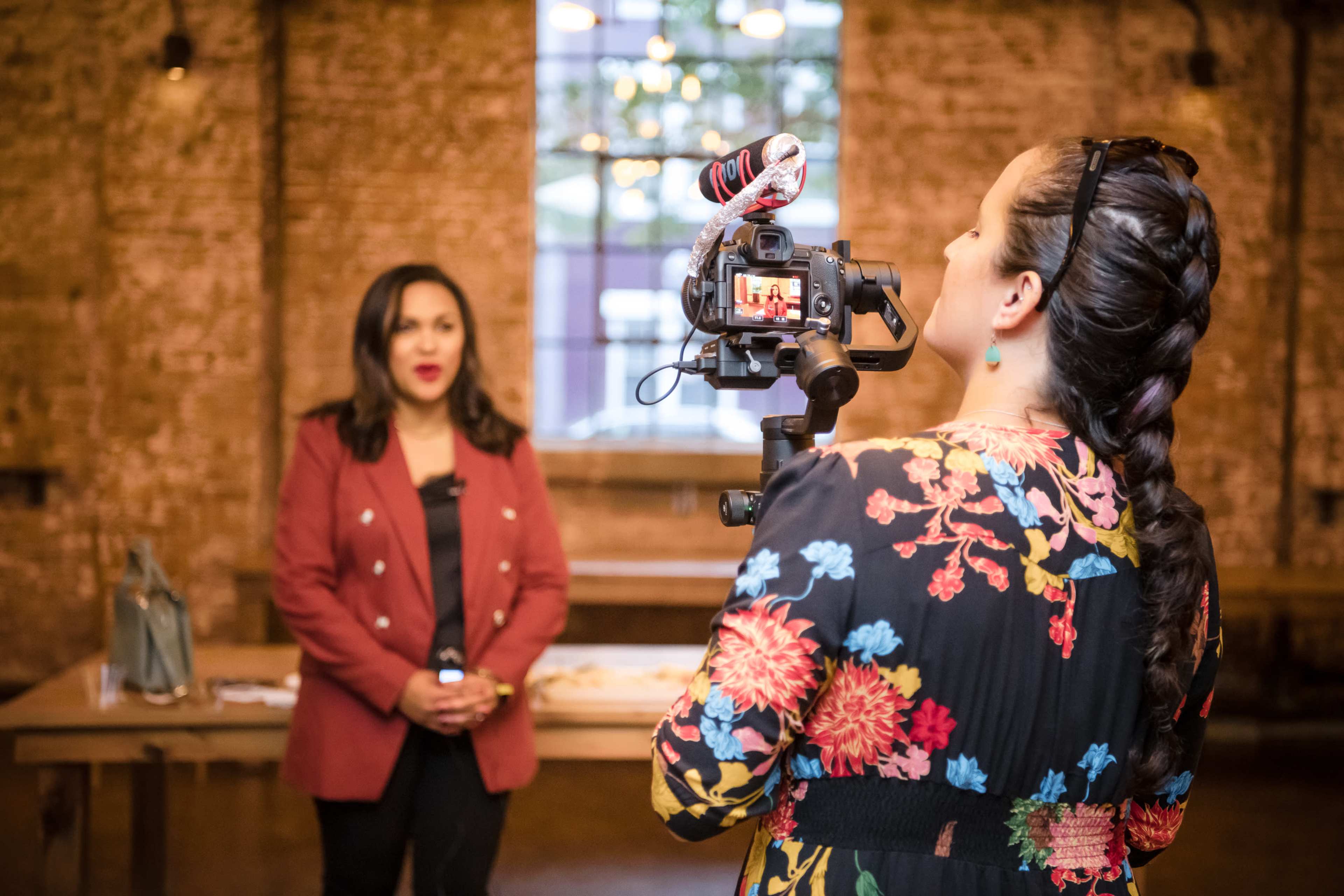 A woman is being filmed while standing at a table in a brick-walled room, as another woman operates the camera.