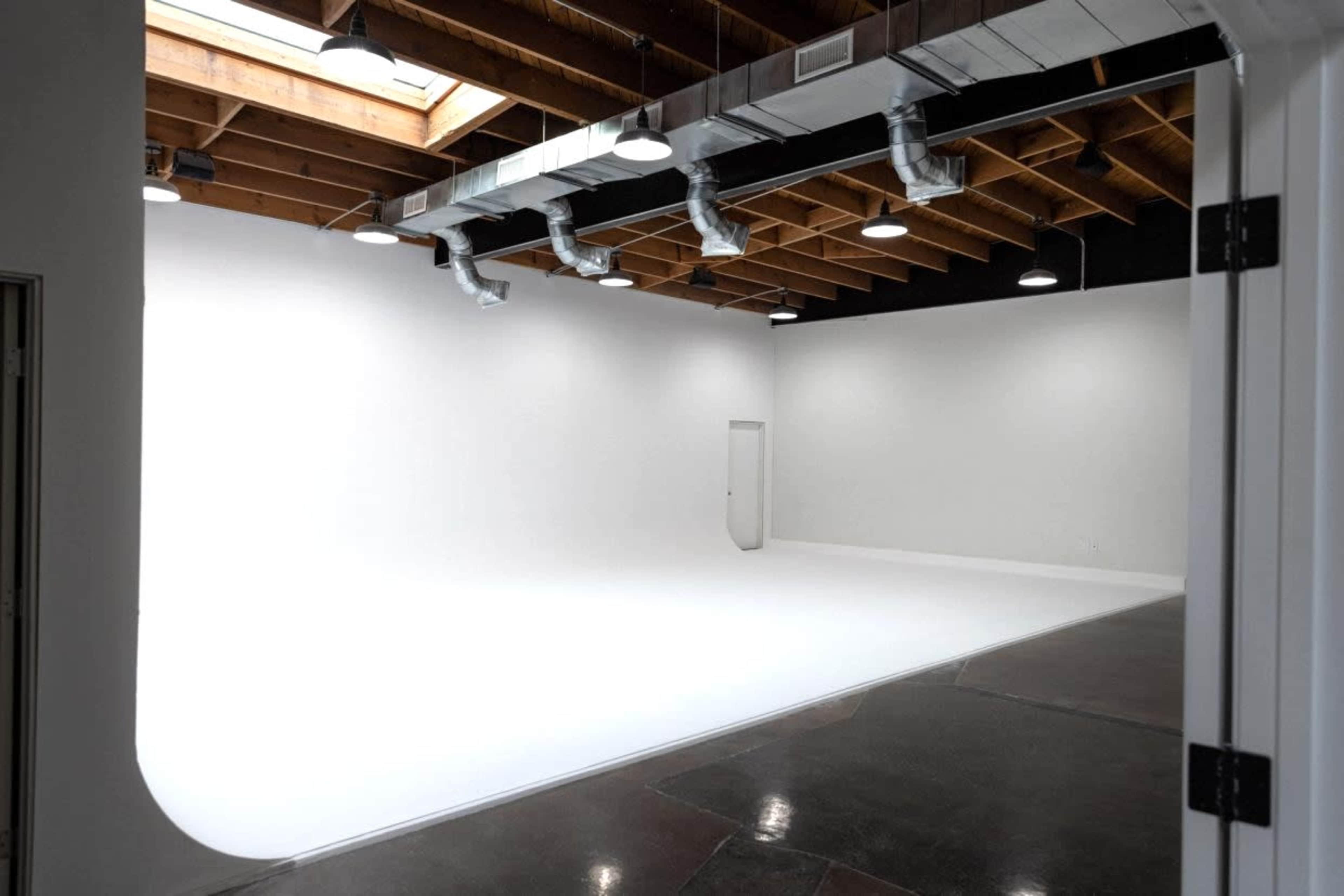 The image shows a large, empty studio space with white walls and a seamless white floor, illuminated by overhead lights.