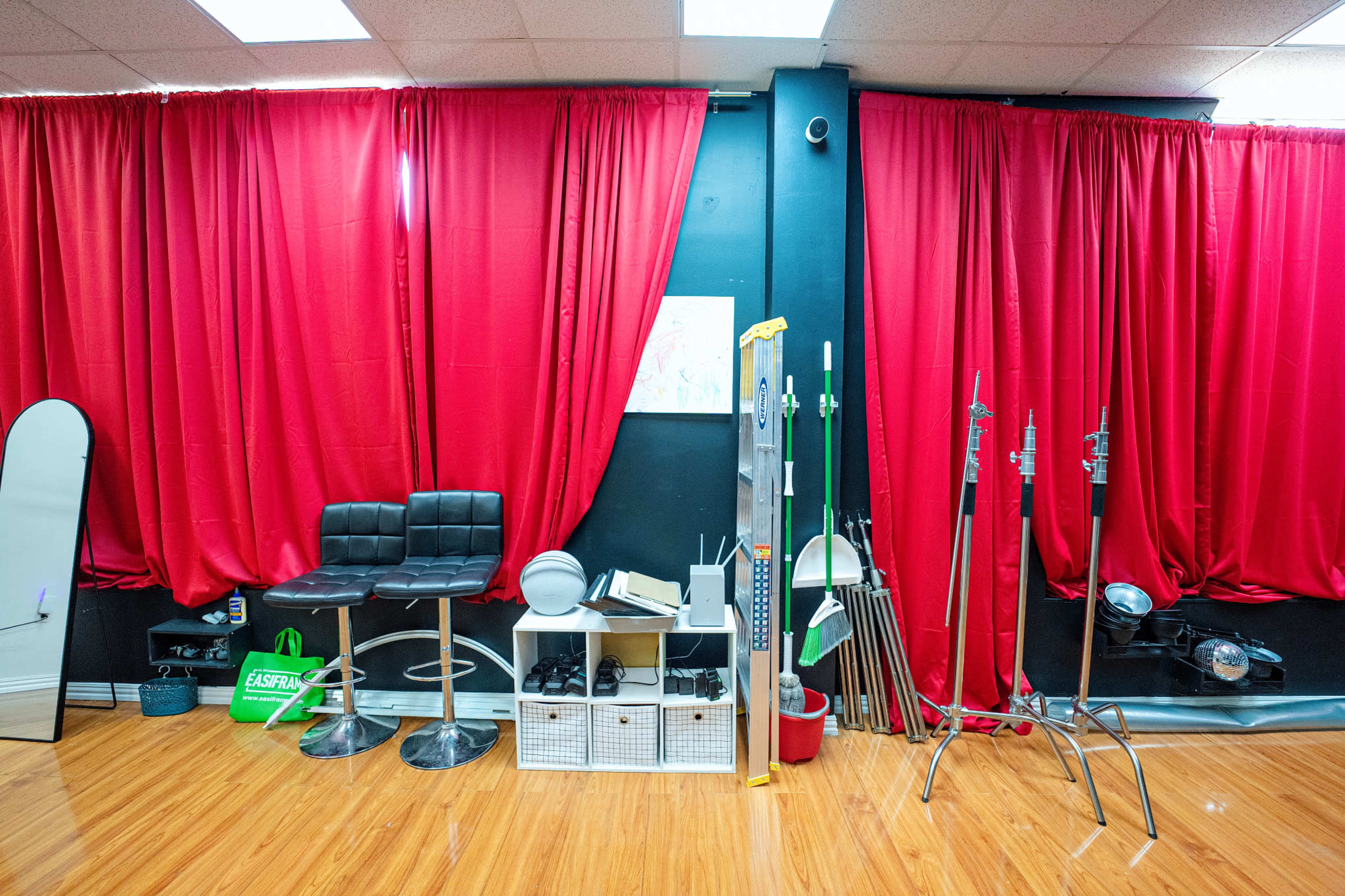 The image shows a corner of a room with red curtains, a pair of black stools, various equipment including stands and a mirror, and wooden flooring.