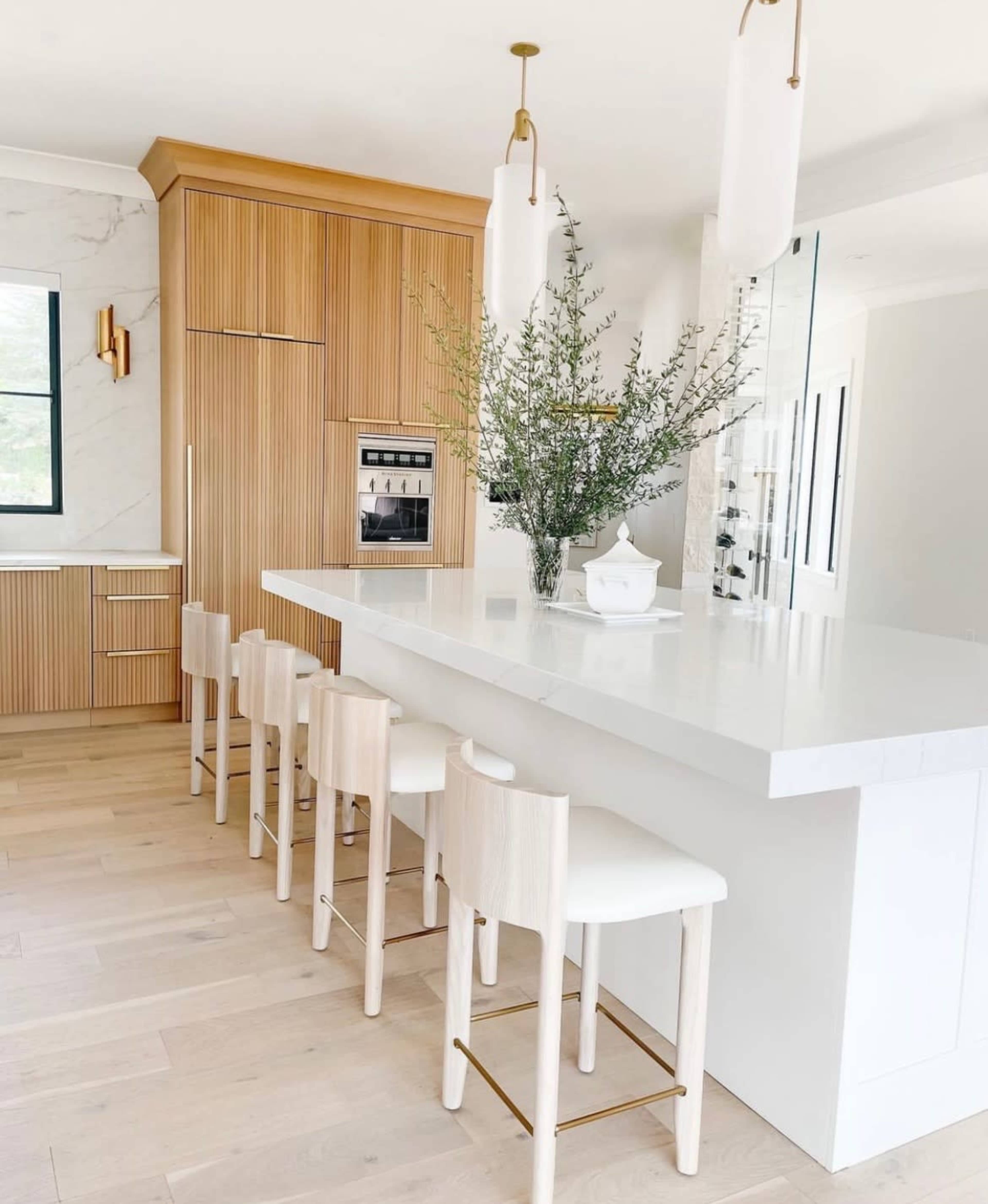 A modern kitchen features a white countertop island with wooden bar stools, complemented by cabinetry with vertical wood slats and a decorative plant centerpiece.