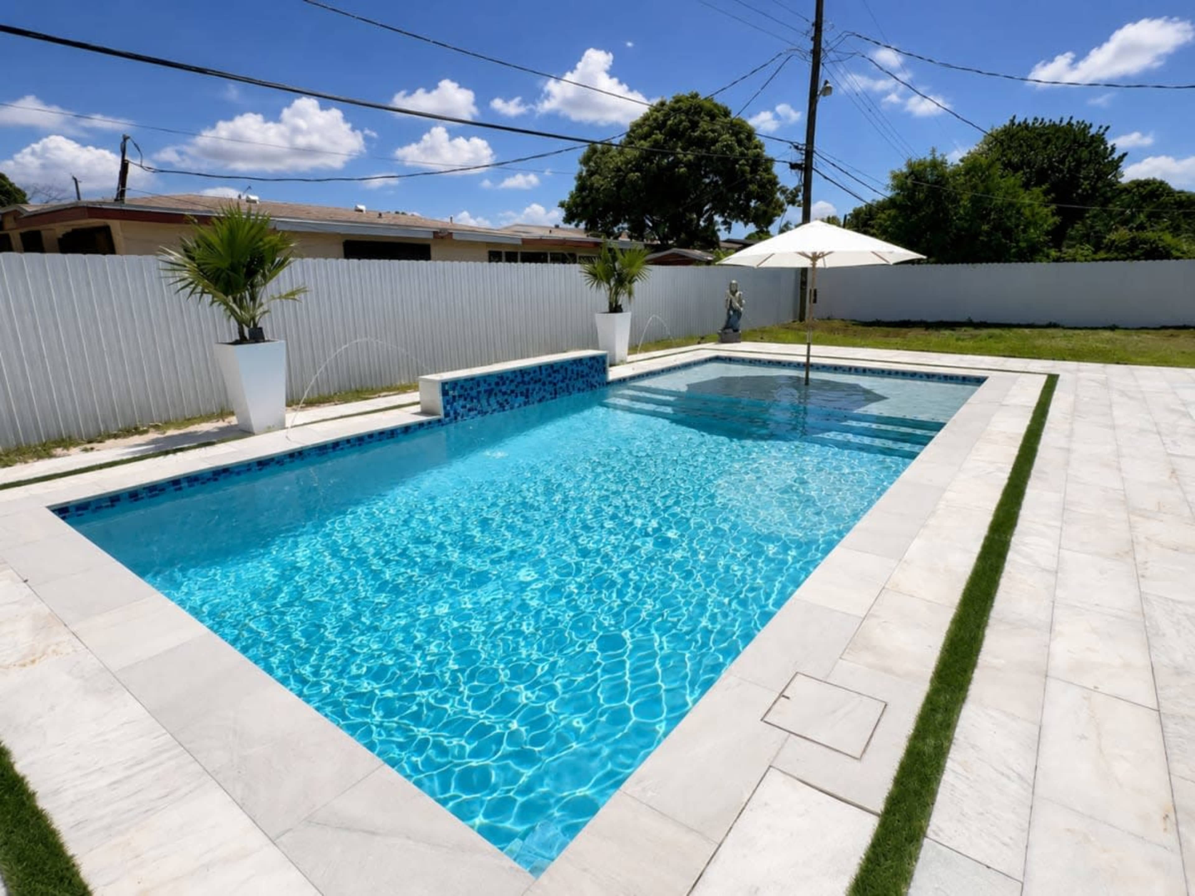 The image shows a rectangular swimming pool surrounded by a white fence, featuring a patio with potted plants and a shaded area provided by an umbrella.