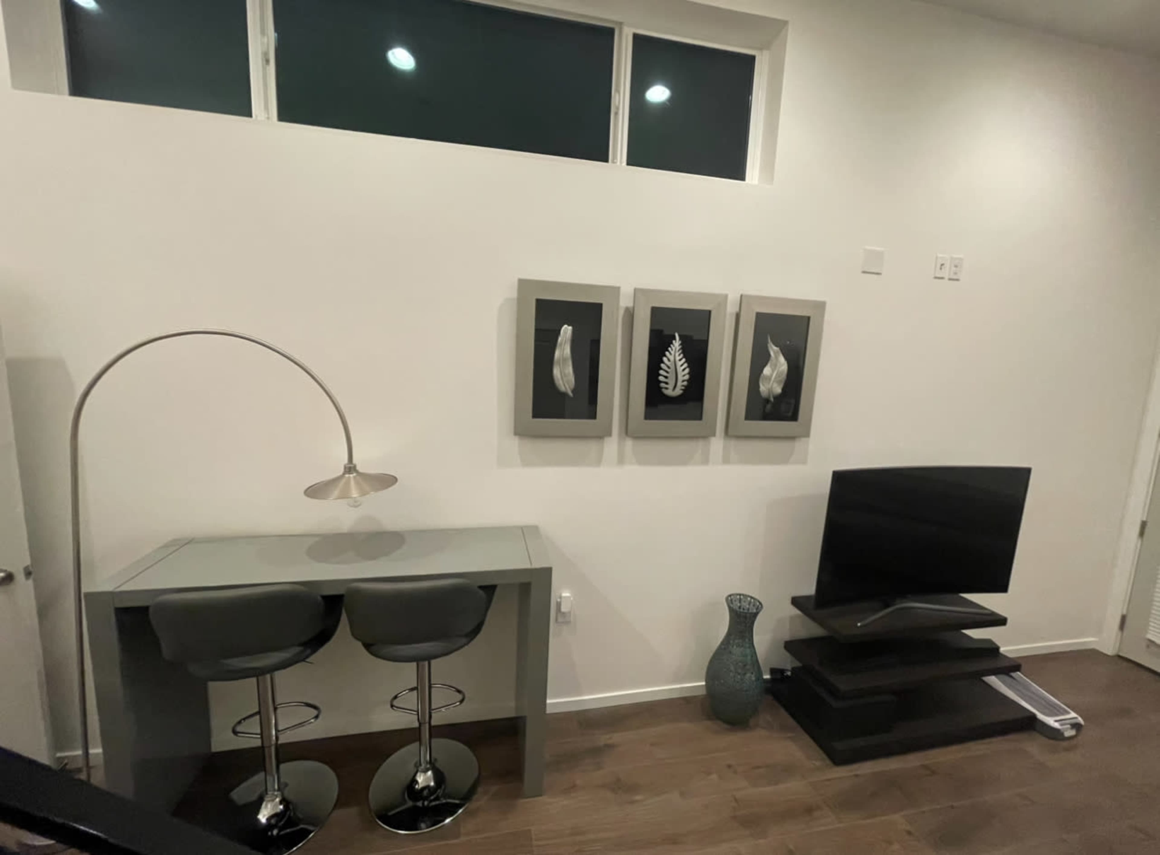 A small modern room features a glass desk with two stools, three framed artworks on the wall, and a television on a black stand accompanied by a decorative vase.