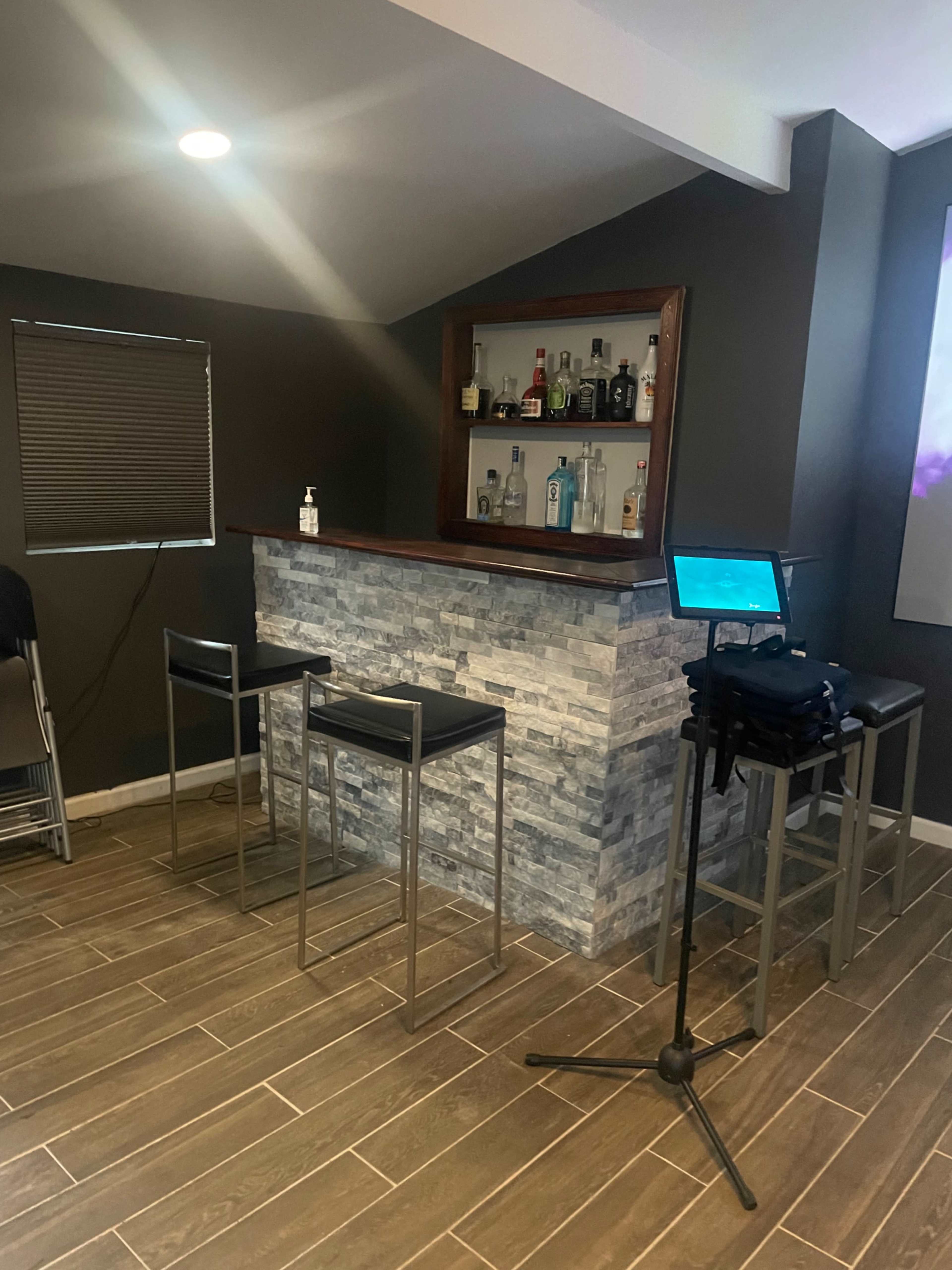 A home bar with a stone facade and shelves displaying various bottles, accompanied by two high stools and a small table with a laptop.