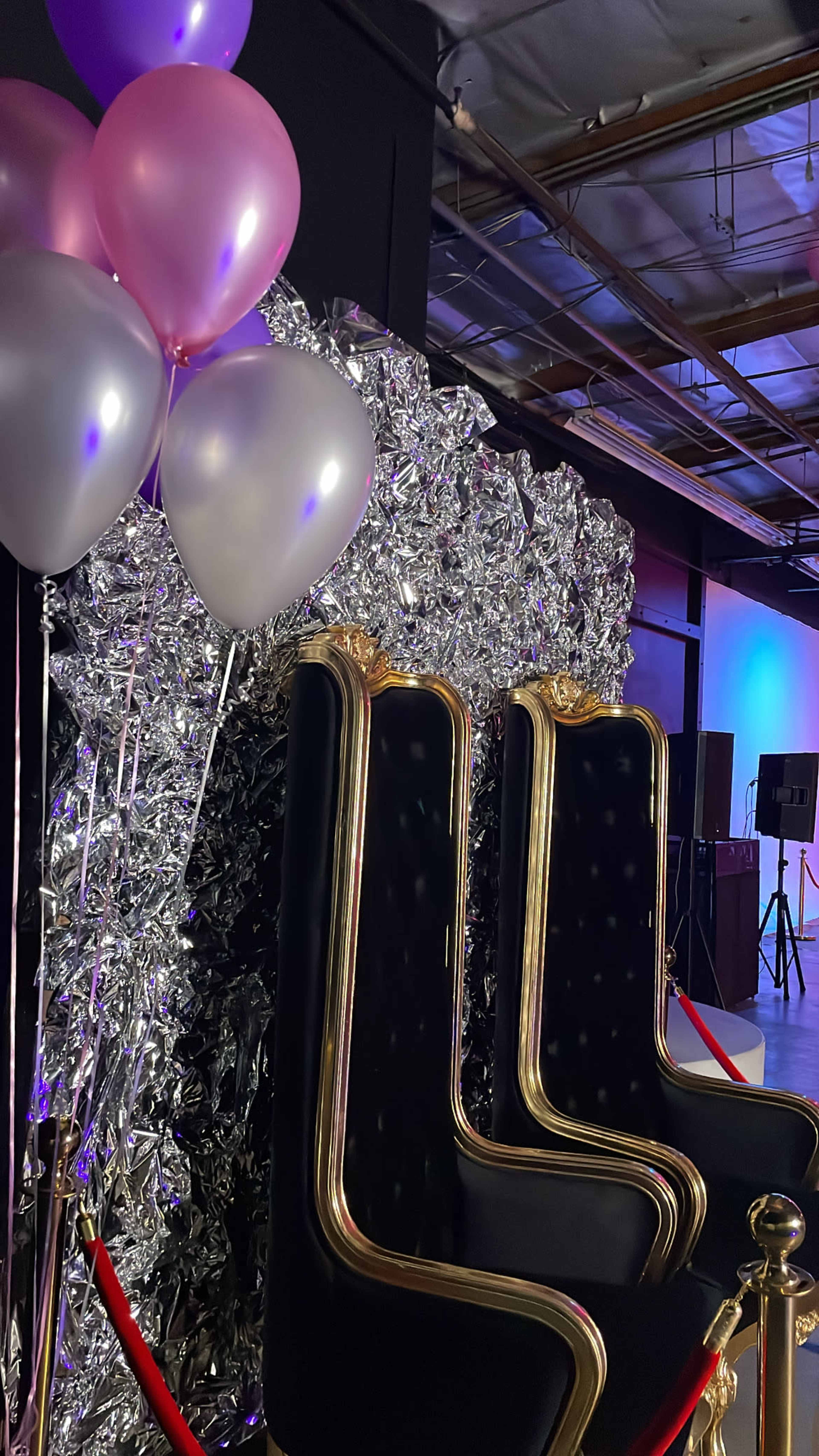 The image shows two ornate black chairs with golden accents in front of a shiny silver backdrop, accompanied by purple and silver balloons.