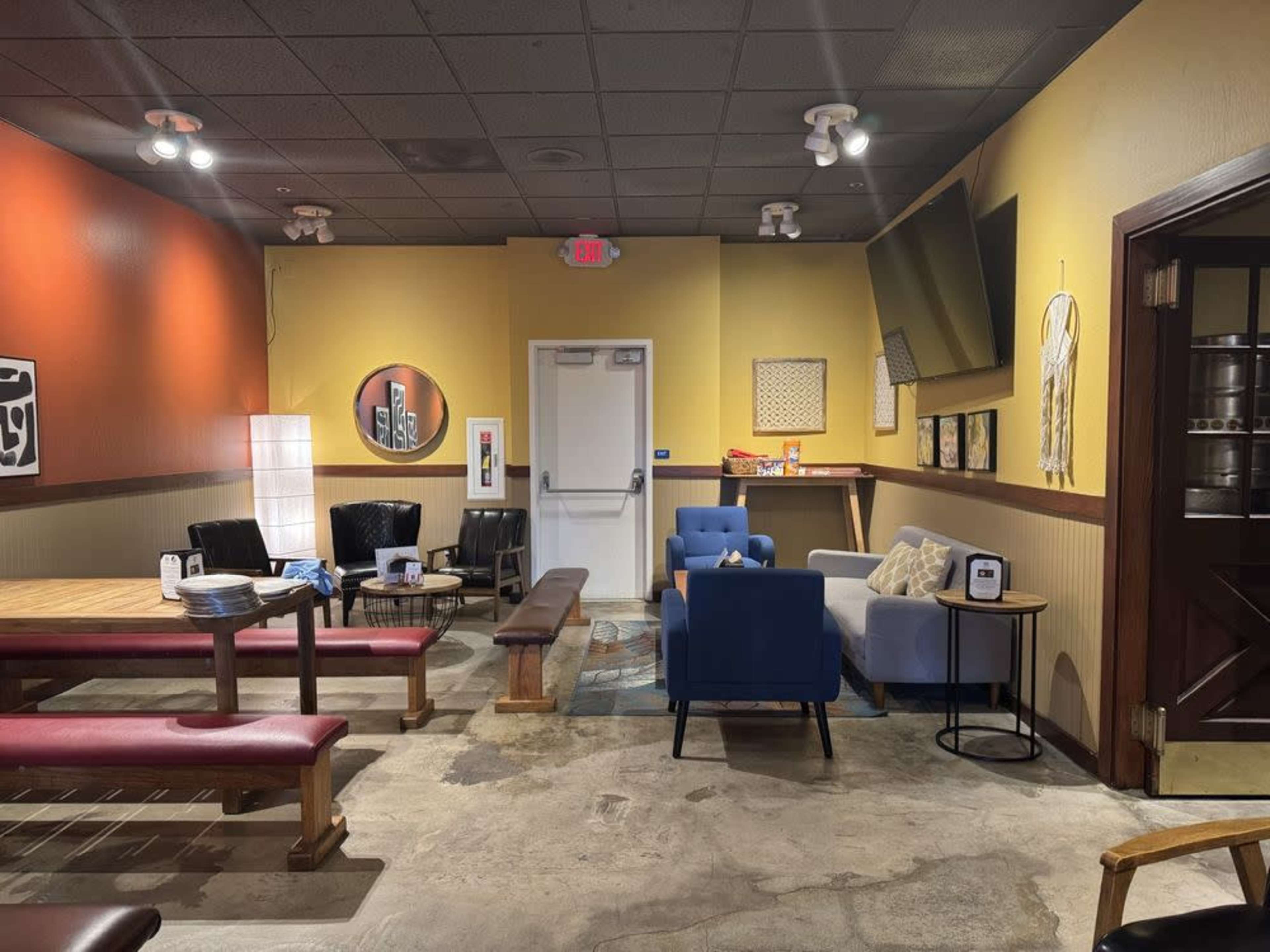 The image shows a cozy lounge area with seating including sofas and chairs, a large wall-mounted television, and colorful walls, arranged in a familiar layout.