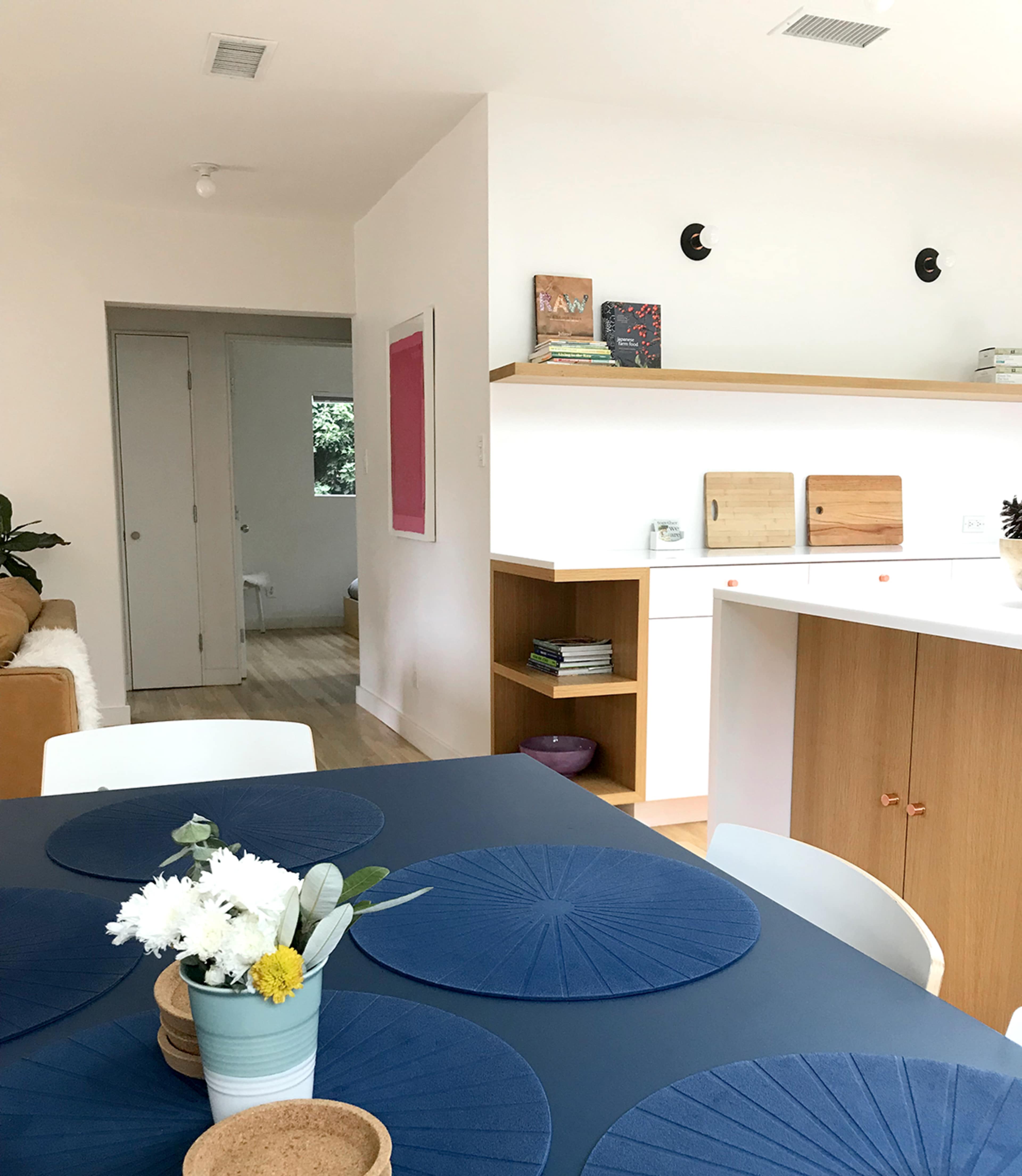 A modern dining area features a table with blue placemats, surrounded by white chairs, and a view into a well-lit kitchen and adjoining room with neutral decor.