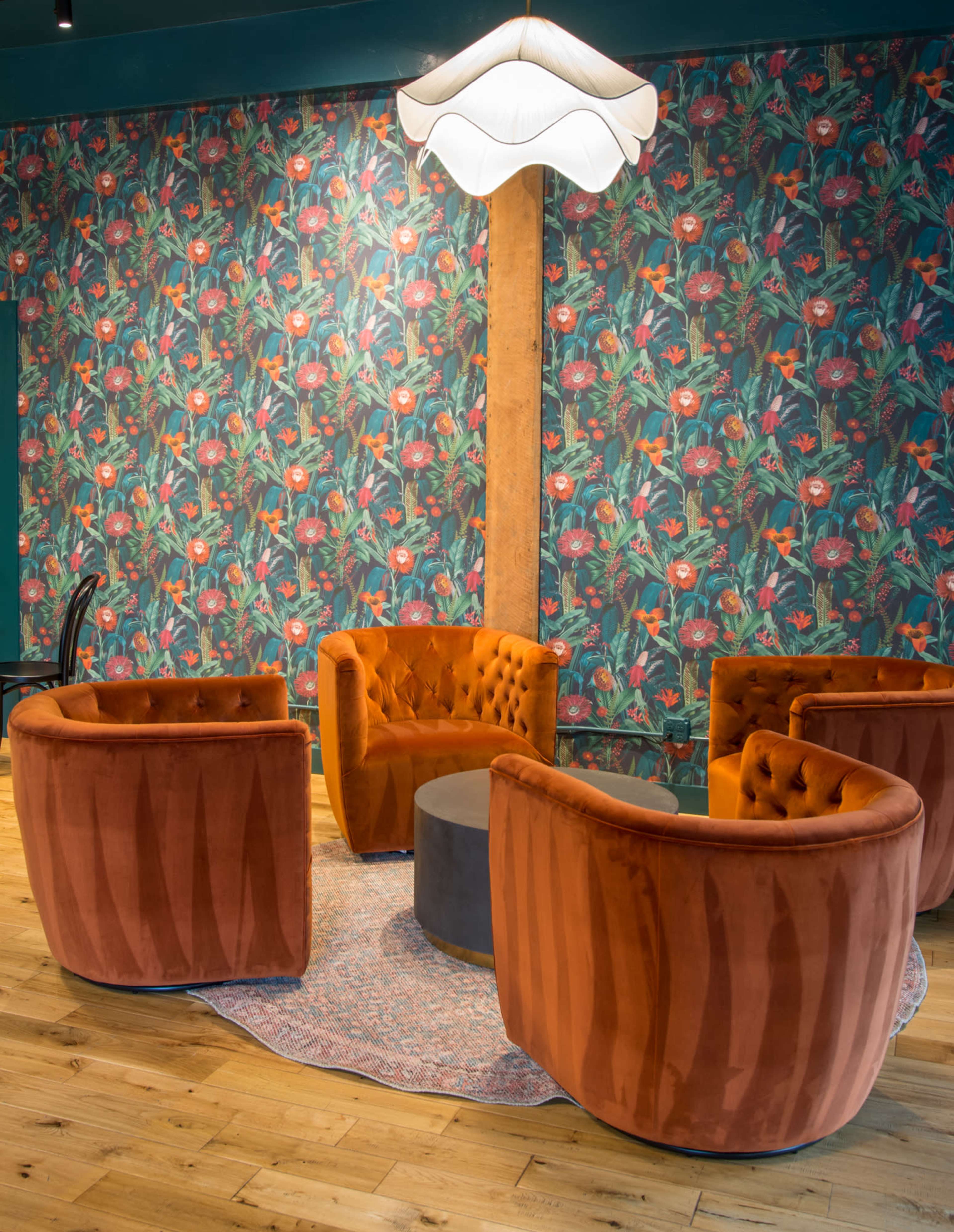 A cozy seating area features four orange velvet chairs arranged around a round table on a patterned rug, set against a floral wallpaper backdrop.