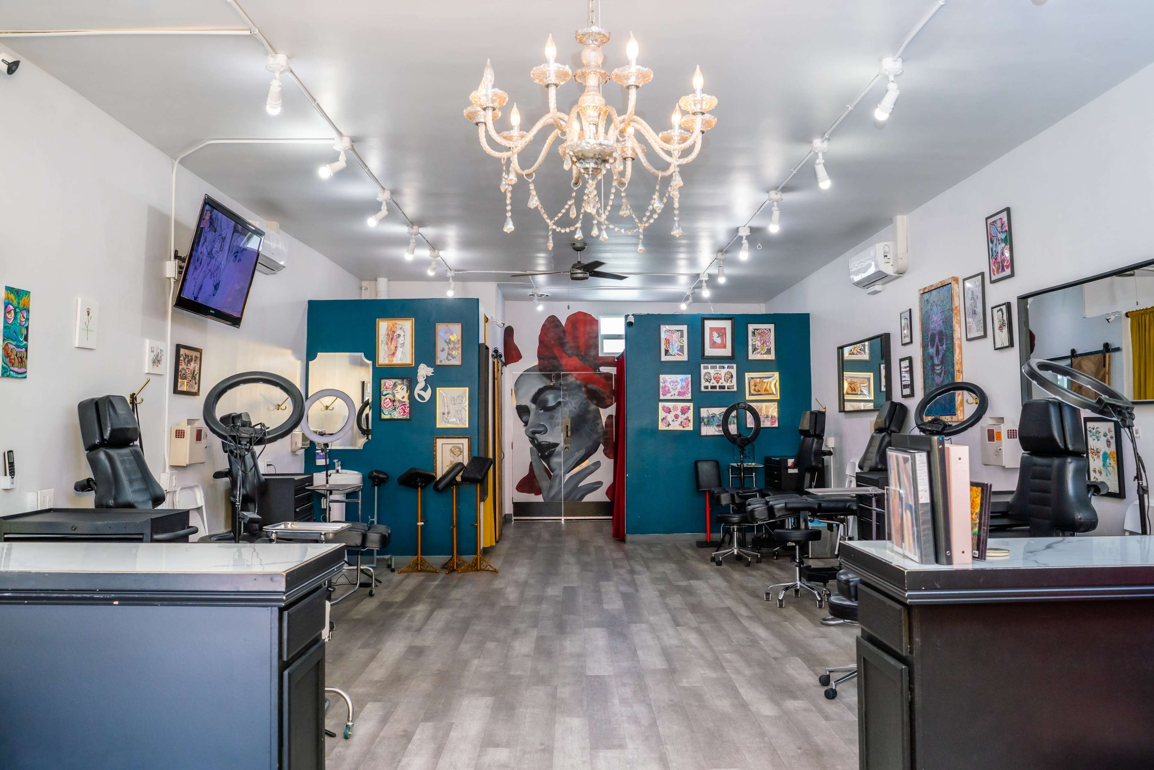 The image shows a tattoo studio featuring multiple workstations, a chandelier, and walls adorned with framed artwork.