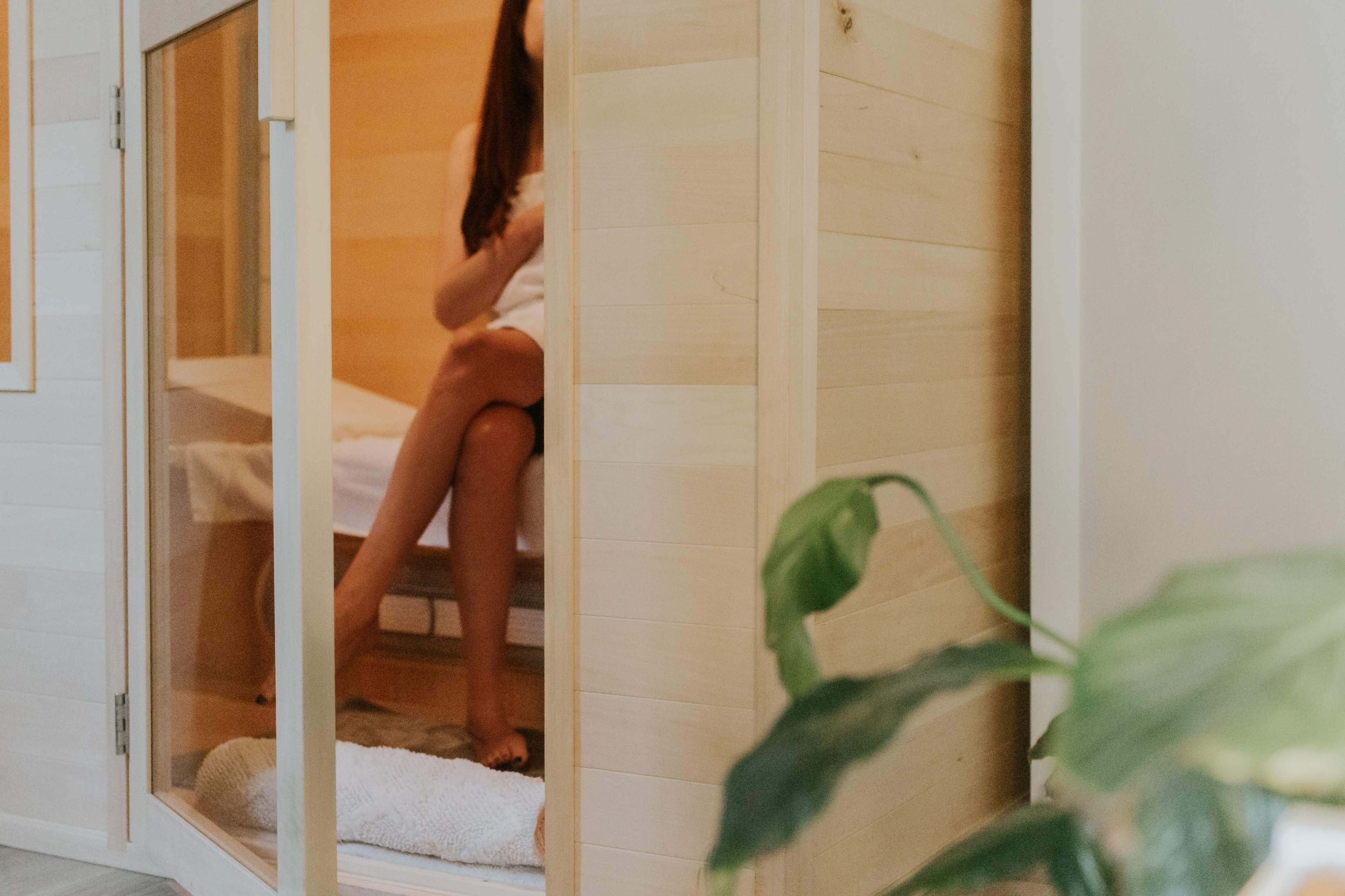A person sits inside a wooden sauna, partially obscured, with a plant in the foreground.
