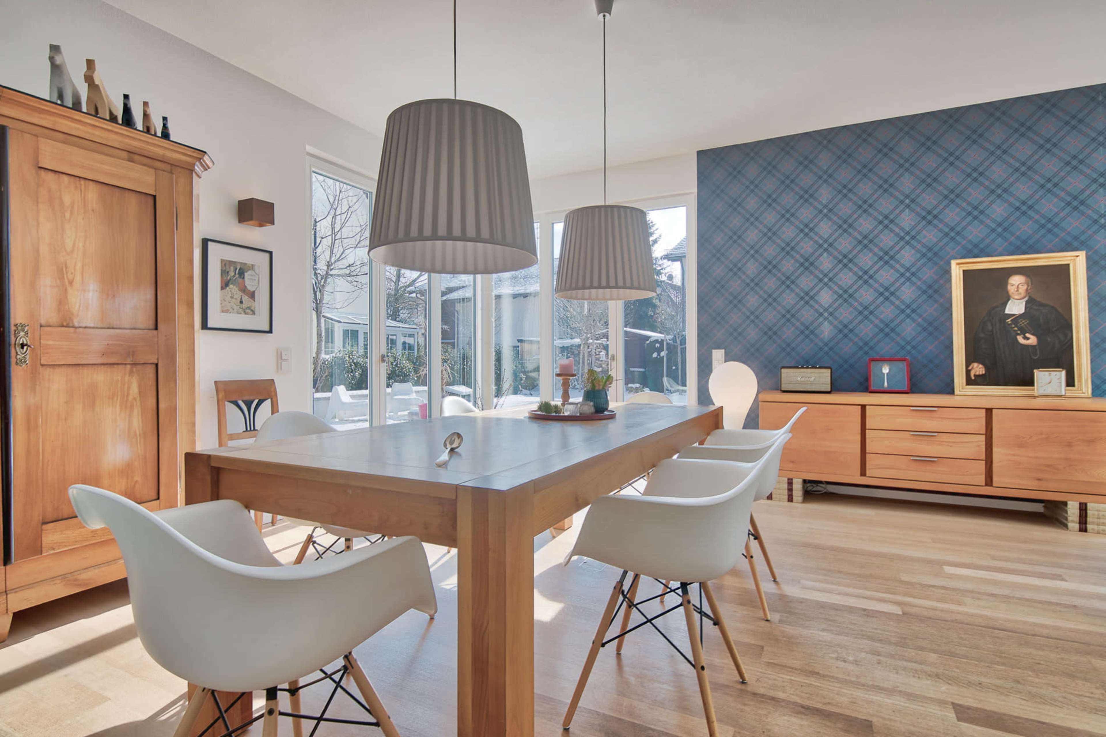 The image features a modern dining room with a wooden table, white chairs, and a large window overlooking an outdoor space, complemented by a blue plaid accent wall and various decorative items.