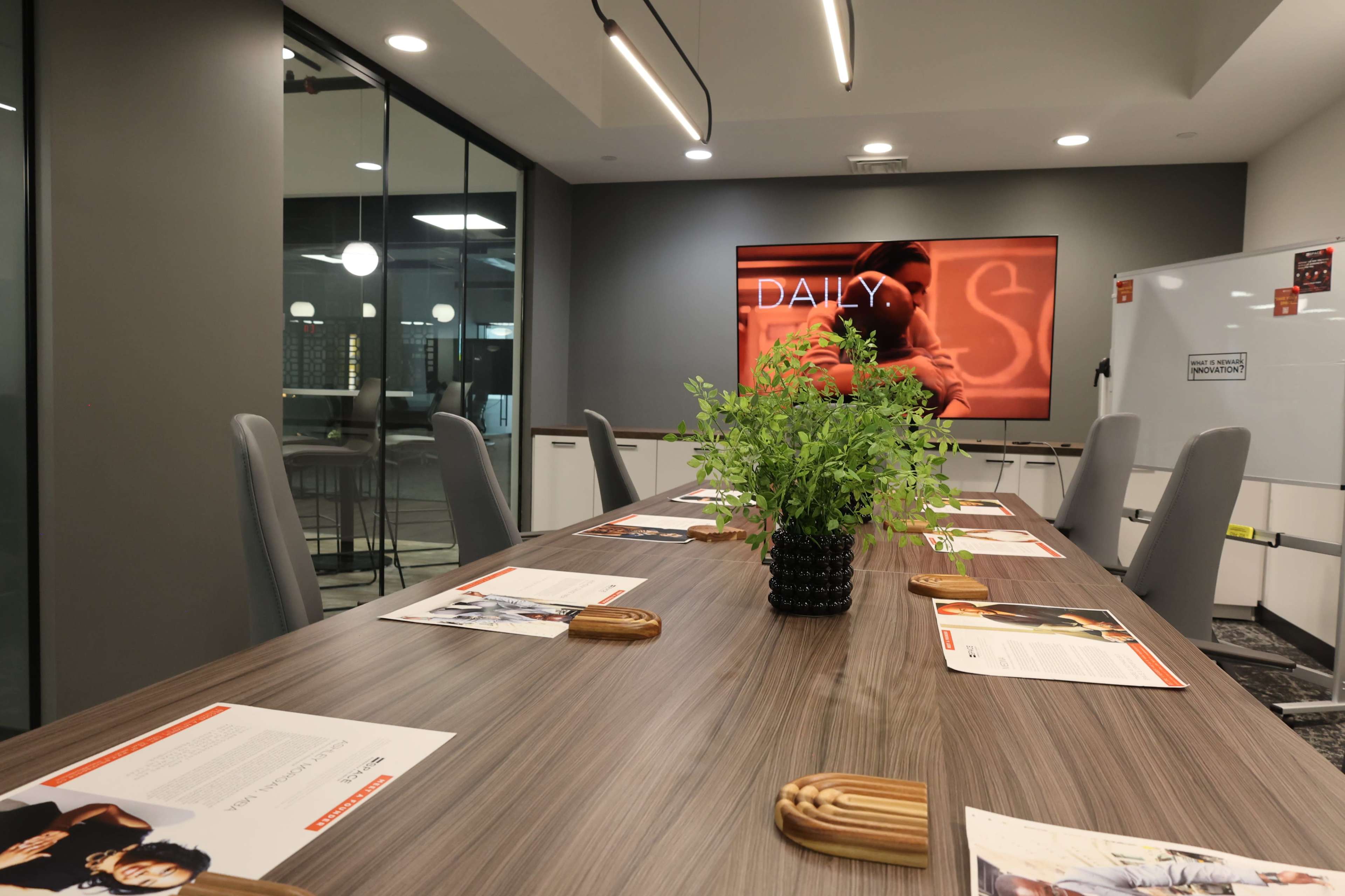 A modern conference room features a long wooden table surrounded by gray chairs, with a large screen displaying the word "DAILY" and a potted plant as the centerpiece.
