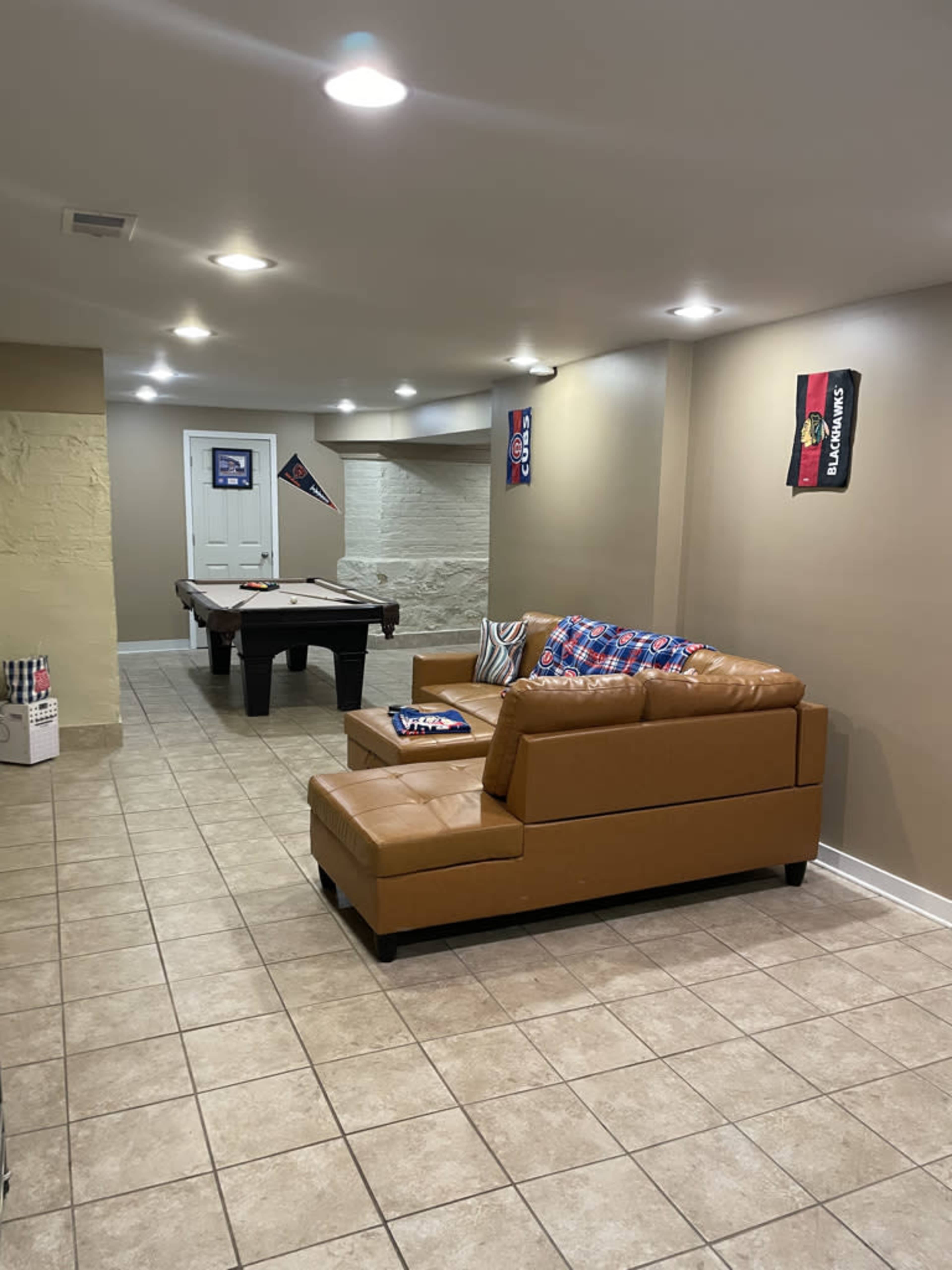 A basement recreation room featuring a tan sectional sofa, a pool table, and wall decorations including sports flags.