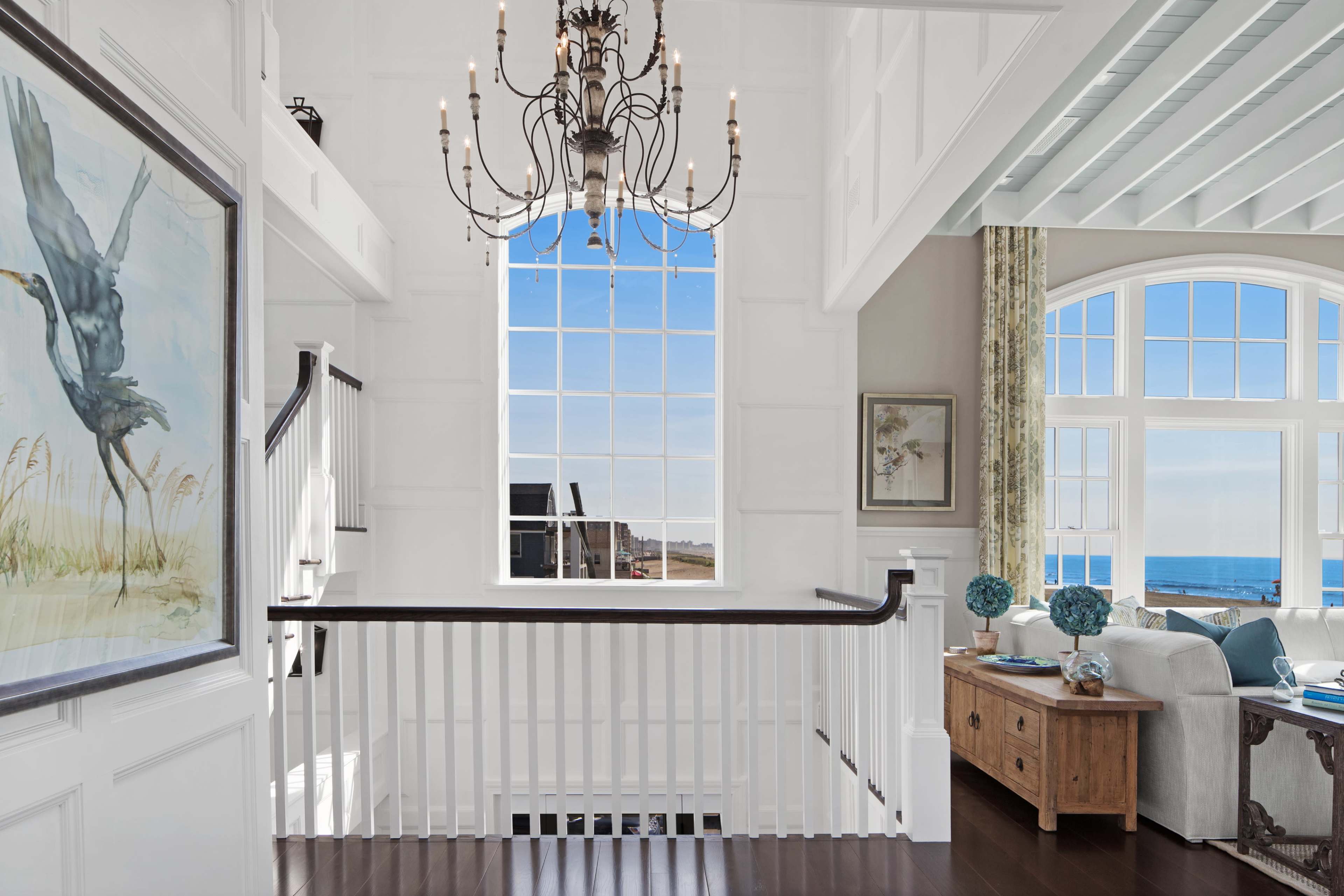A well-lit interior features a large window overlooking the ocean, framed by white walls and a wooden railing, with an art piece of a bird and a decorative table nearby.