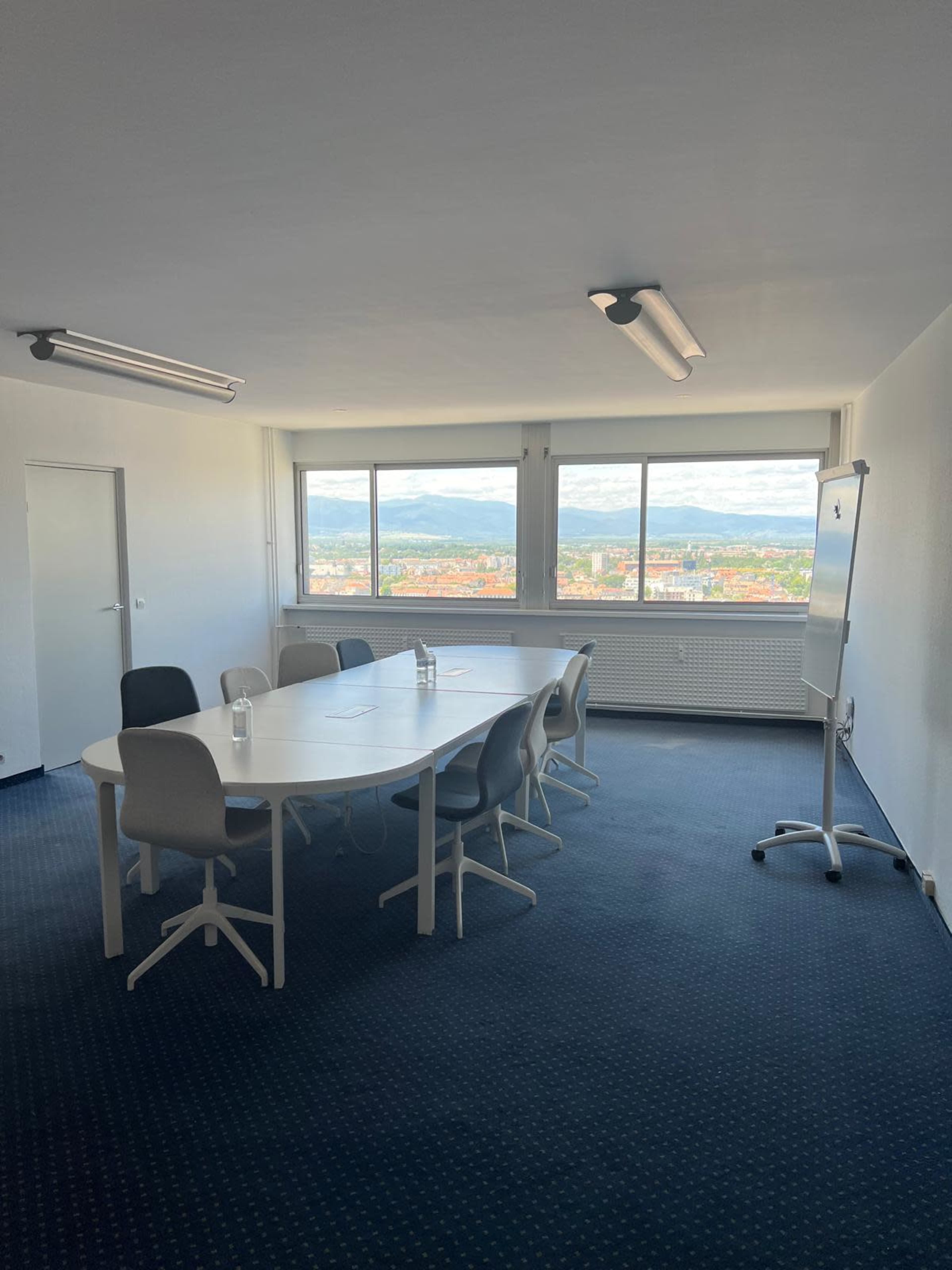 The image shows a modern conference room with a large white table, several chairs, and a view of a cityscape through large windows.