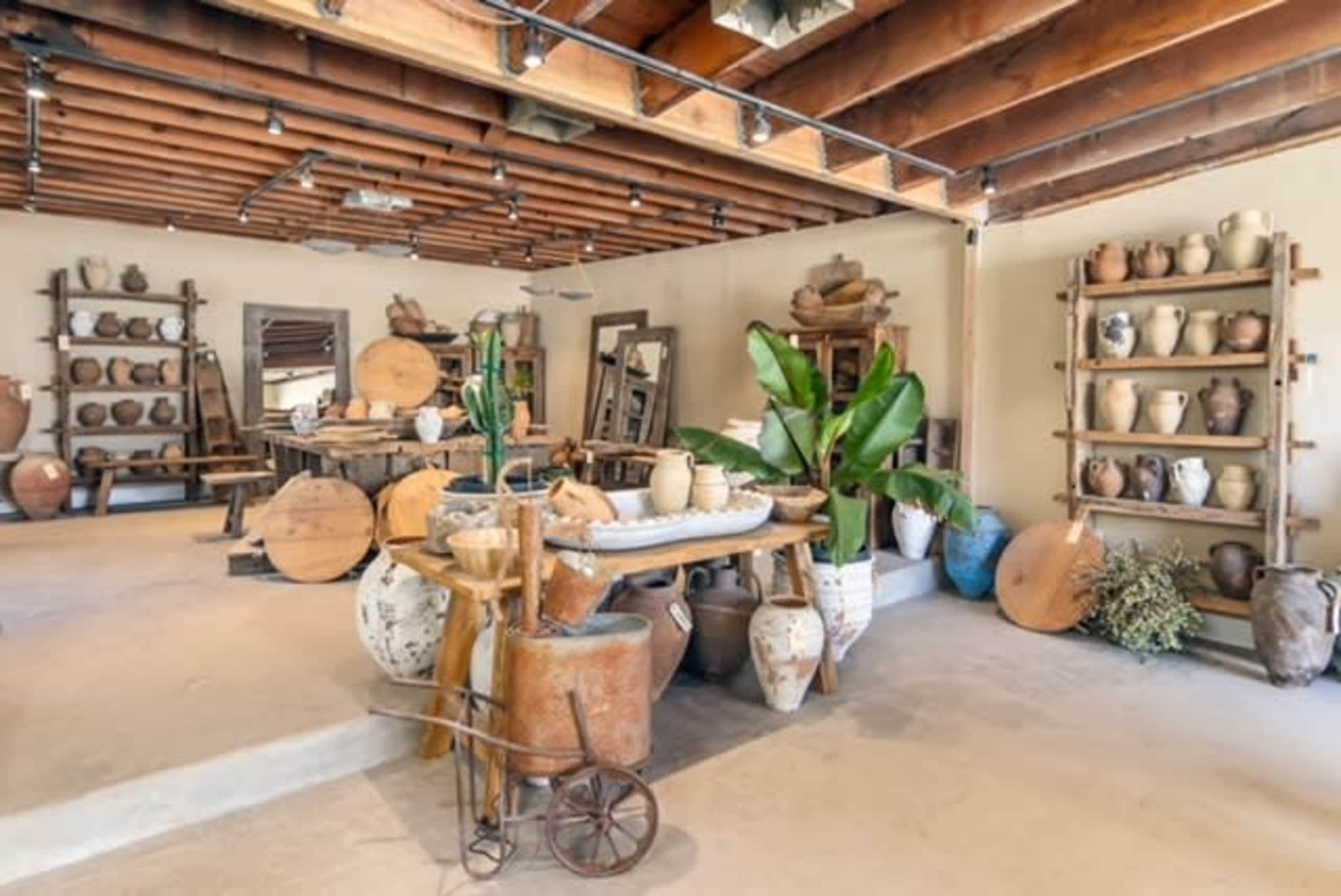 The image shows a spacious interior filled with various pottery pieces, wooden shelves, and decorative plants.
