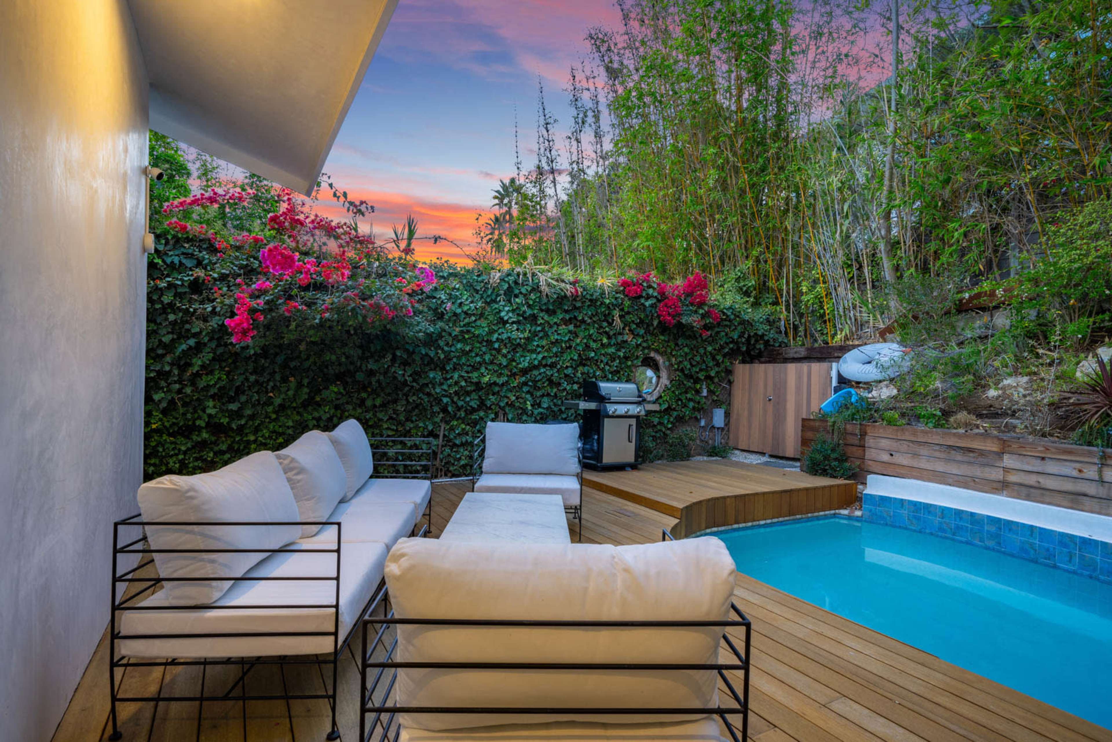 A modern outdoor patio features white sofas and a small swimming pool surrounded by lush greenery and colorful flowers at sunset.