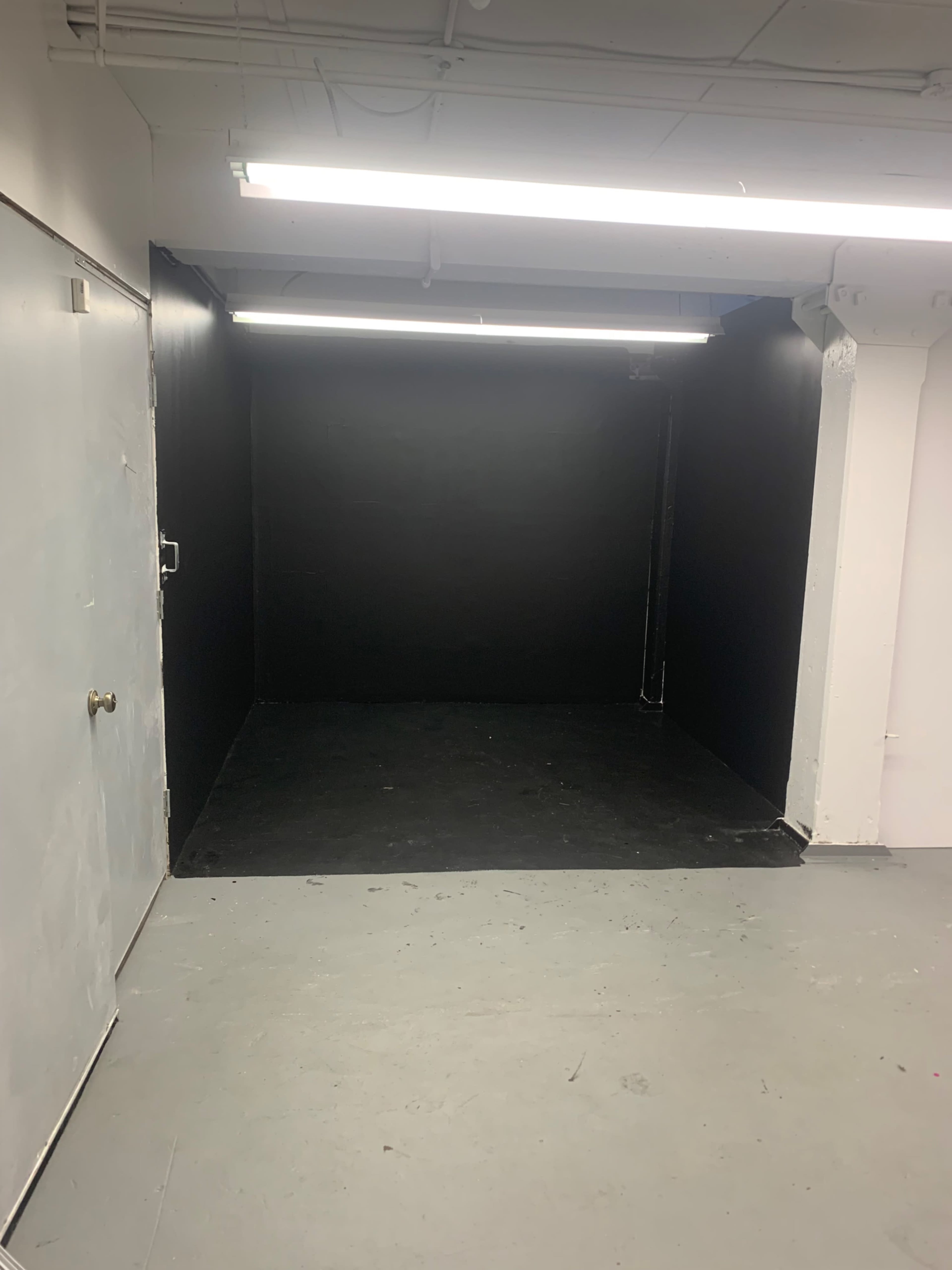 The image shows an empty room with black walls and a black floor, illuminated by fluorescent lights above.