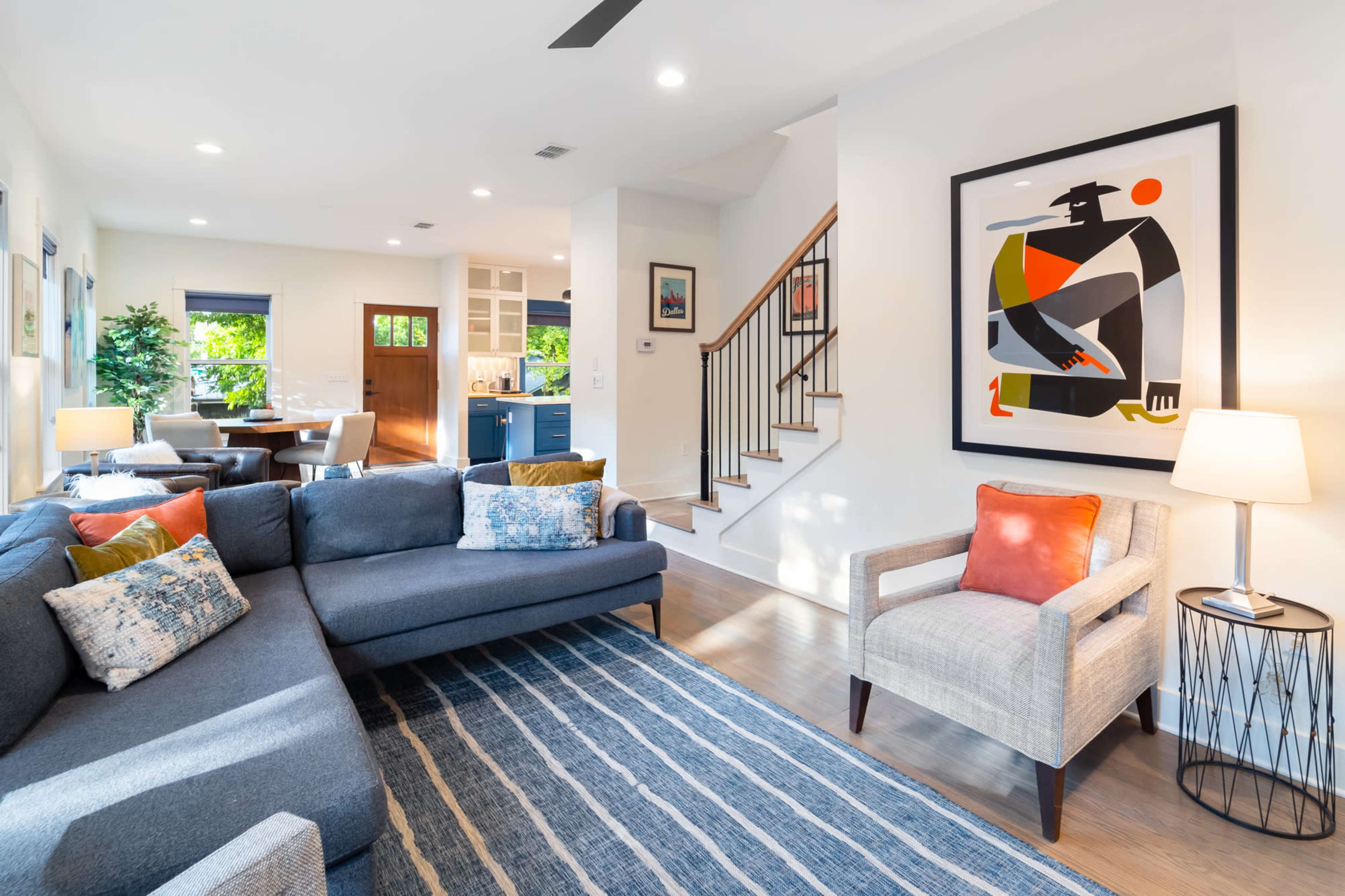 The image shows a modern living room with a grey sectional sofa, a patterned rug, and a staircase leading to an upper level, illuminated by natural light from large windows.