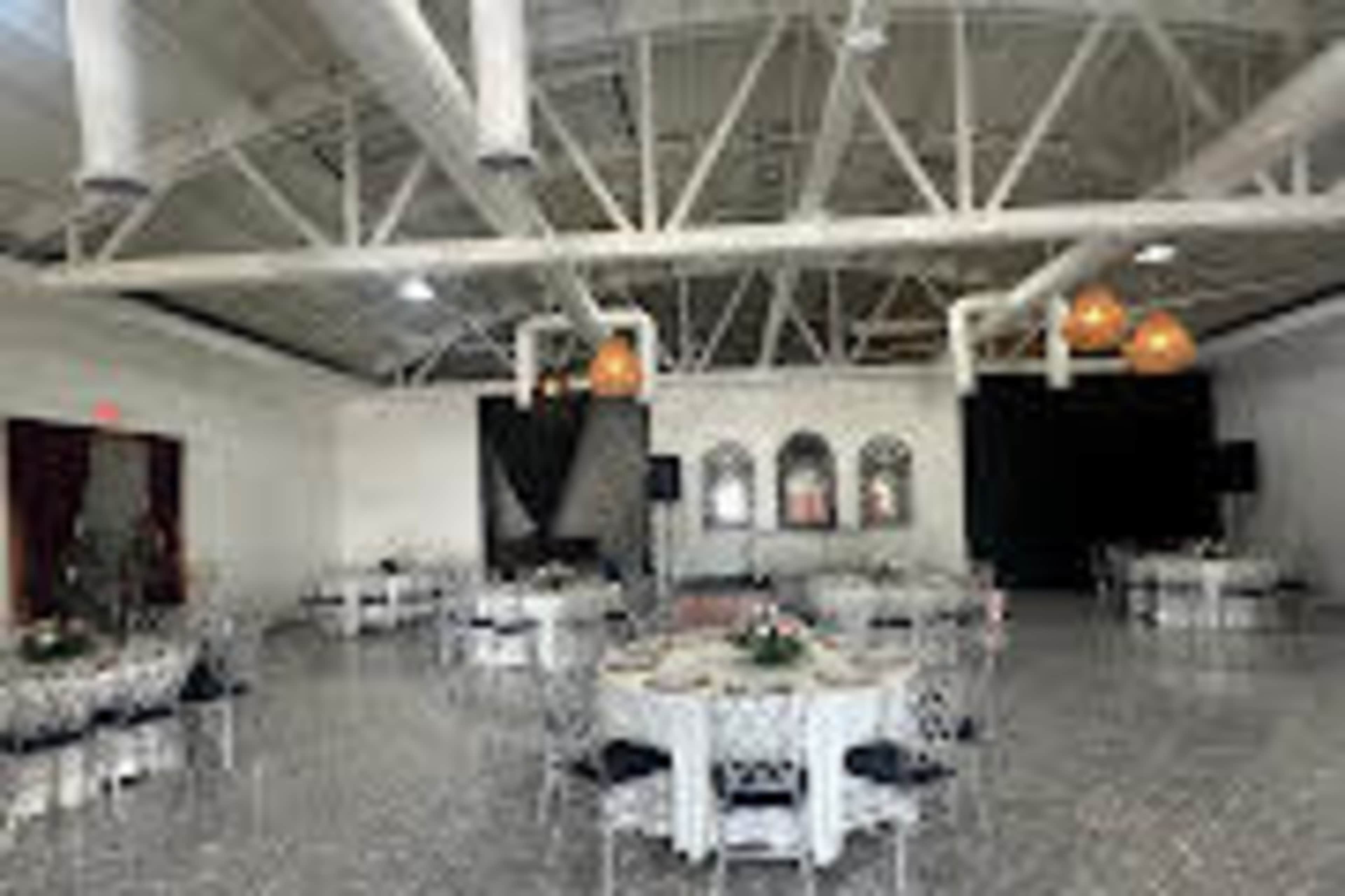 The image shows a spacious event venue with round tables set up, decorated with centerpieces, and large windows on one side.