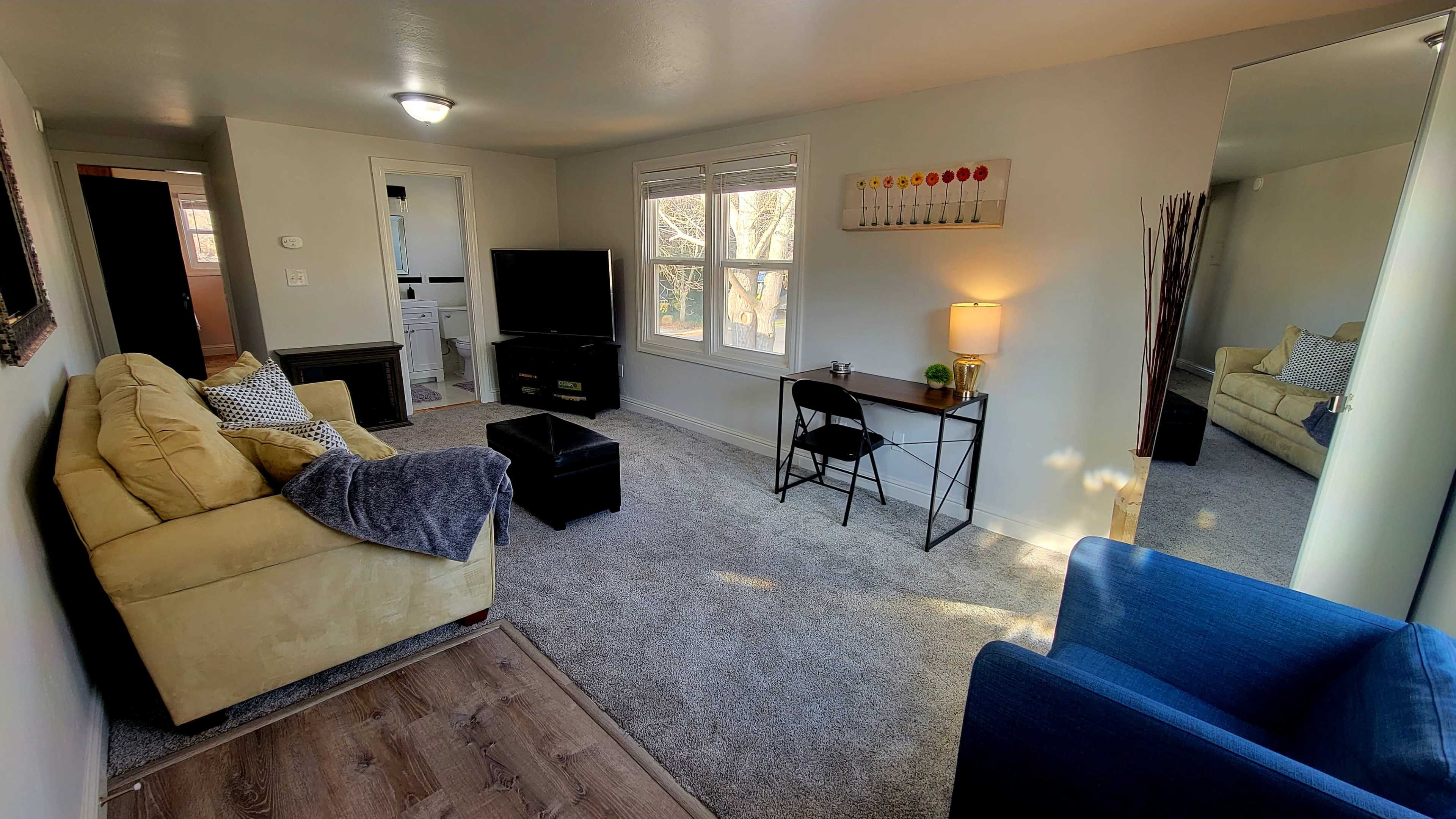 The living room features a yellow sofa, a blue chair, a coffee table, a small desk with a lamp, and a television against a light-colored wall.